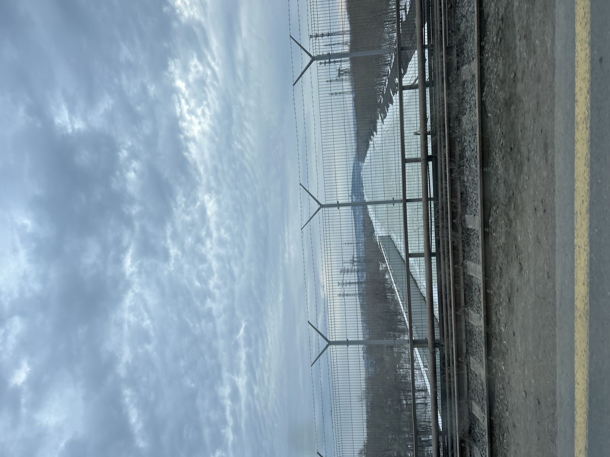 The image depicts a railroad scene in what is likely Chaikovsky, Russia, given the likely location. The foreground shows a section of a train platform edge marked with a yellow safety line. Adjacent to the platform are railroad tracks lined with ballast. A high wire fence runs parallel to the tracks, topped with barbed wire. Beyond the fence, a vista of electrical towers and distant hills is visible under an overcast sky. The scene conveys a sense of an industrial or transportation zone.