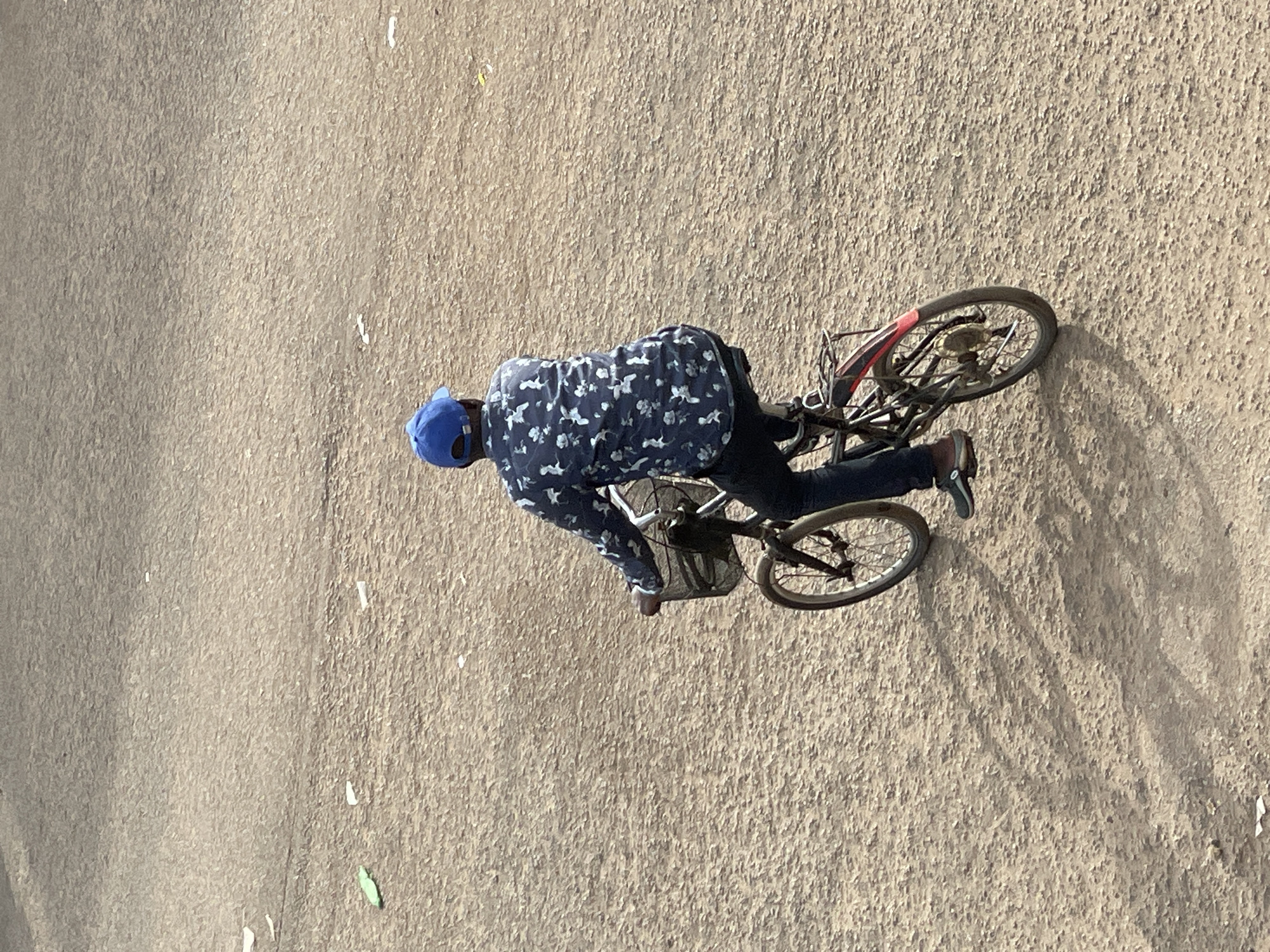 A person wearing a blue cap and a patterned long-sleeved shirt is riding a bicycle on a paved surface. The bicycle is dark-colored with red accents on the wheels and has a basket on the front. The person is leaning forward, gripping the handlebars. The ground appears to be a light brown, rough texture, possibly asphalt or dirt. The sun is casting shadows, suggesting it is daytime, likely late afternoon or early morning due to the length of the shadows. The scene is outdoors with no other people or objects clearly visible. The image is taken from a high angle, looking down on the cyclist. There is no visible text in the image.