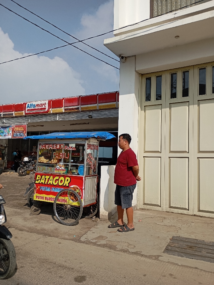 The image depicts an outdoor commercial scene in Situwangi, Indonesia, under a bright, partly cloudy sky. In the center, a red, white, and blue mobile food cart, labeled "BATAGOR" and "PUTRA FAMILI," is positioned on a paved sidewalk. The cart features glass display cases containing food items and is covered by a blue fabric awning. A bicycle wheel leans against the front of the cart. To the right of the cart, an adult male wearing a maroon short-sleeved shirt, dark shorts, and dark sandals stands with his hands clasped behind his back, facing towards the right. He is located in front of a large, cream-colored building with tall, multi-paneled doors. A white security camera is mounted on the upper corner of this building. To the left of the food cart, a red and white Alfamart convenience store with its distinct roof structure and signage is visible. Multiple motorcycles are parked or partially visible in the vicinity of the Alfamart and the foreground. Overhead power lines traverse the upper portion of the frame.