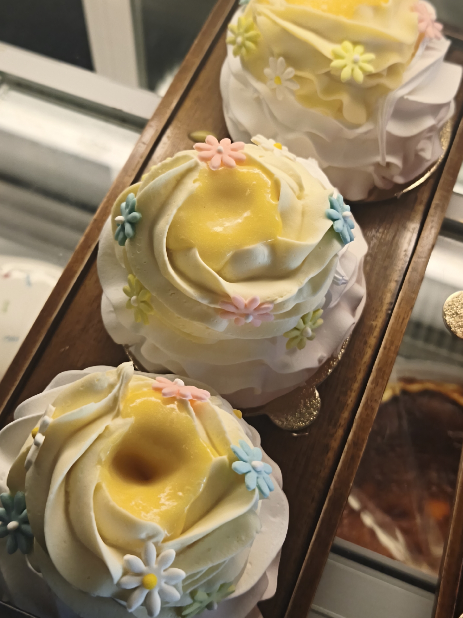 Three cream-frosted cupcakes with yellow filling and decorative candy flowers are arranged on a wooden tray. The cupcakes are individually presented in white paper liners and sit on what appear to be gold bases. The tray itself is dark wood. In the background, a display case is visible with a dark-colored dessert, possibly a pie or tart, to the right. The lighting is bright, and the overall scene suggests a bakery or pastry shop. The location is not specified in the image.
