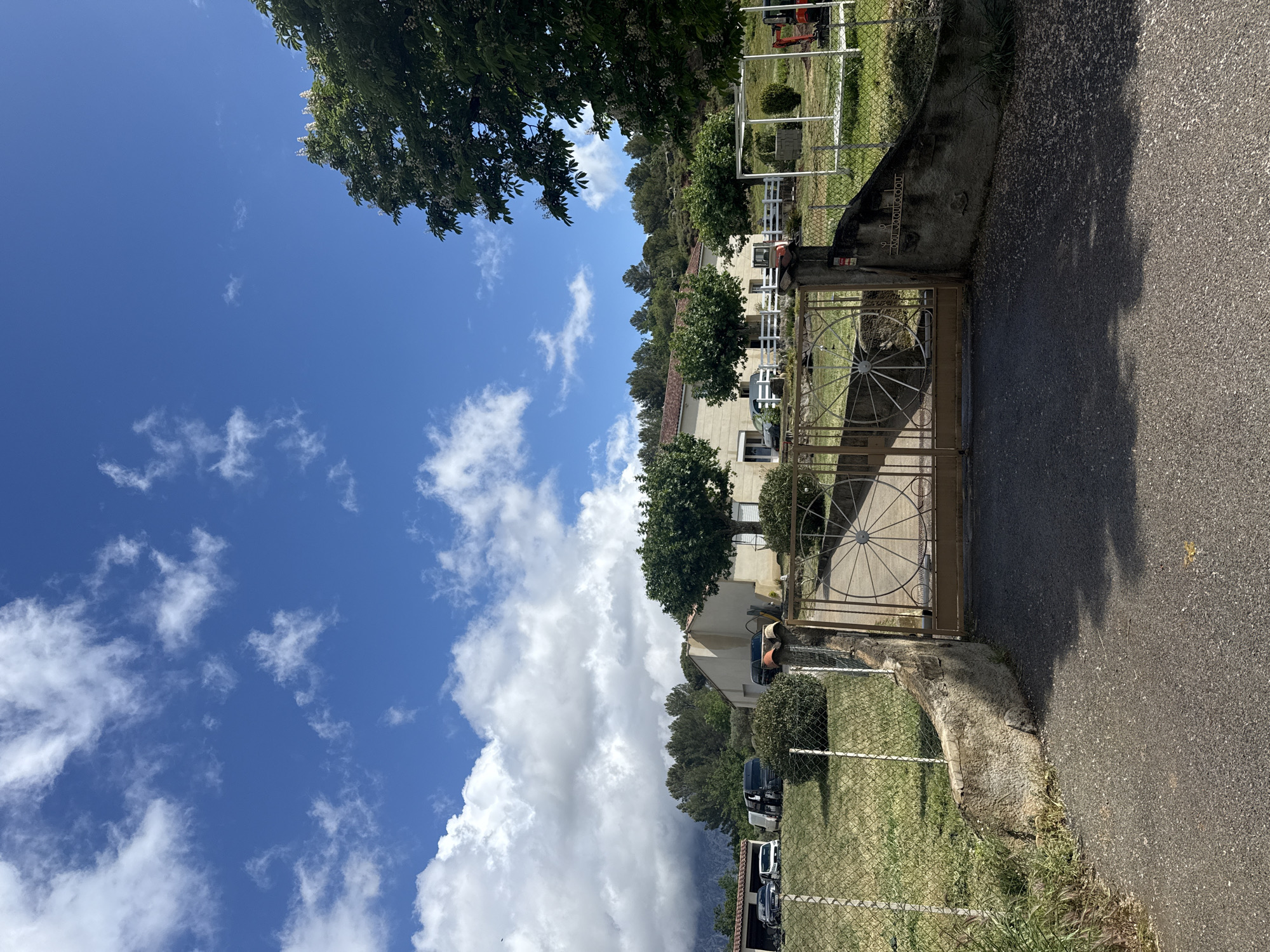 The image depicts an outdoor scene near a gate in Rousset, France. The sky is blue with scattered clouds. The gate is made of metal with a circular design, possibly leading to a residential or commercial property. A paved road is visible in the foreground. In the background, there are buildings, trees, and a fence. The scene is captured in daylight, suggesting a sunny day. Vegetation is present on either side of the gate. There is a concrete or stone structure near the gate. The orientation of the photograph is rotated.