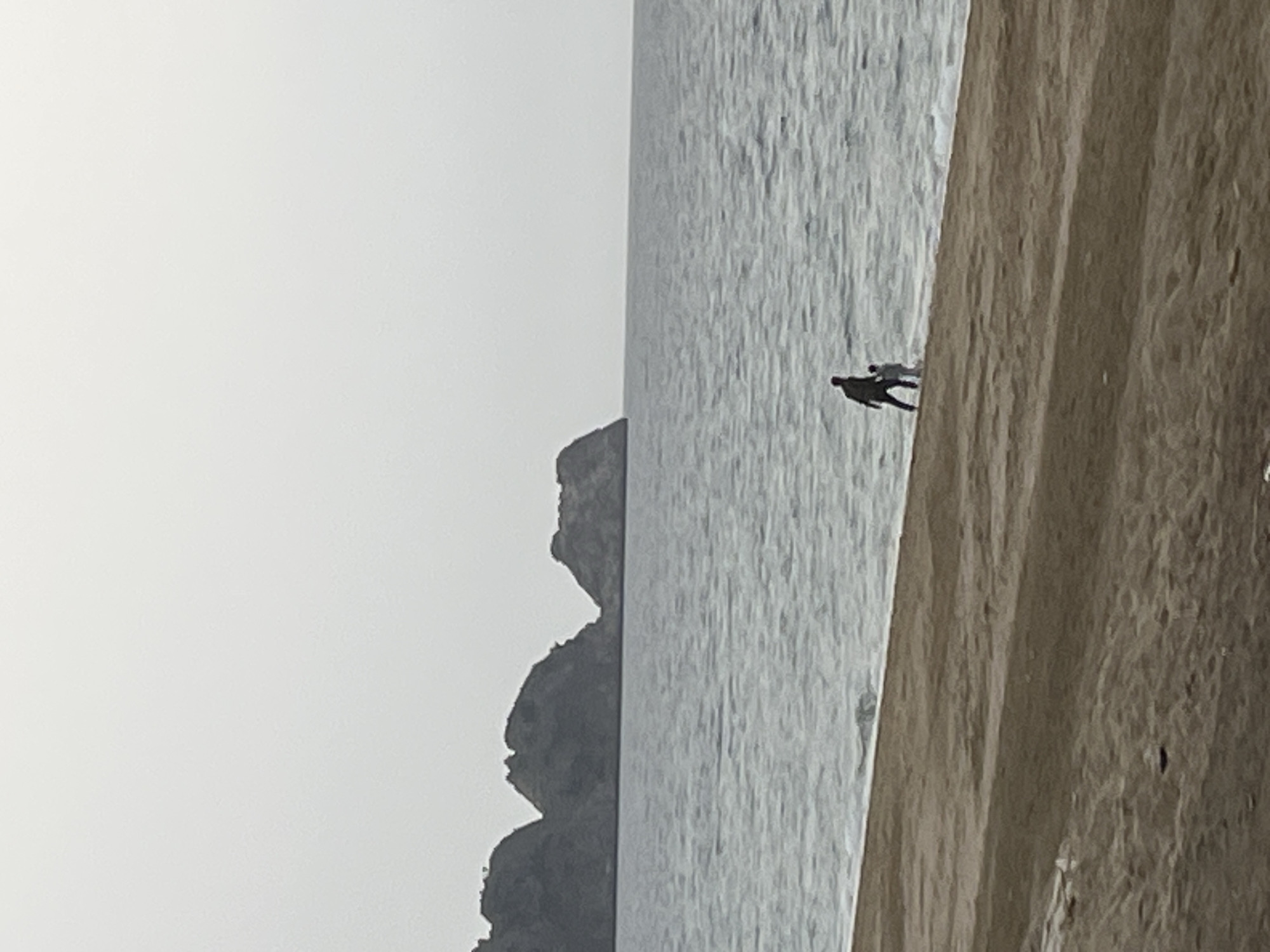 The image shows a beach scene in Alanya, Turkey. The beach is on the right, with light brown sand. The ocean is in the center of the image, with small waves. There is a small mountain or cliff on the left side of the image. There are two people in the water walking along the beach. The sky is overcast and white.