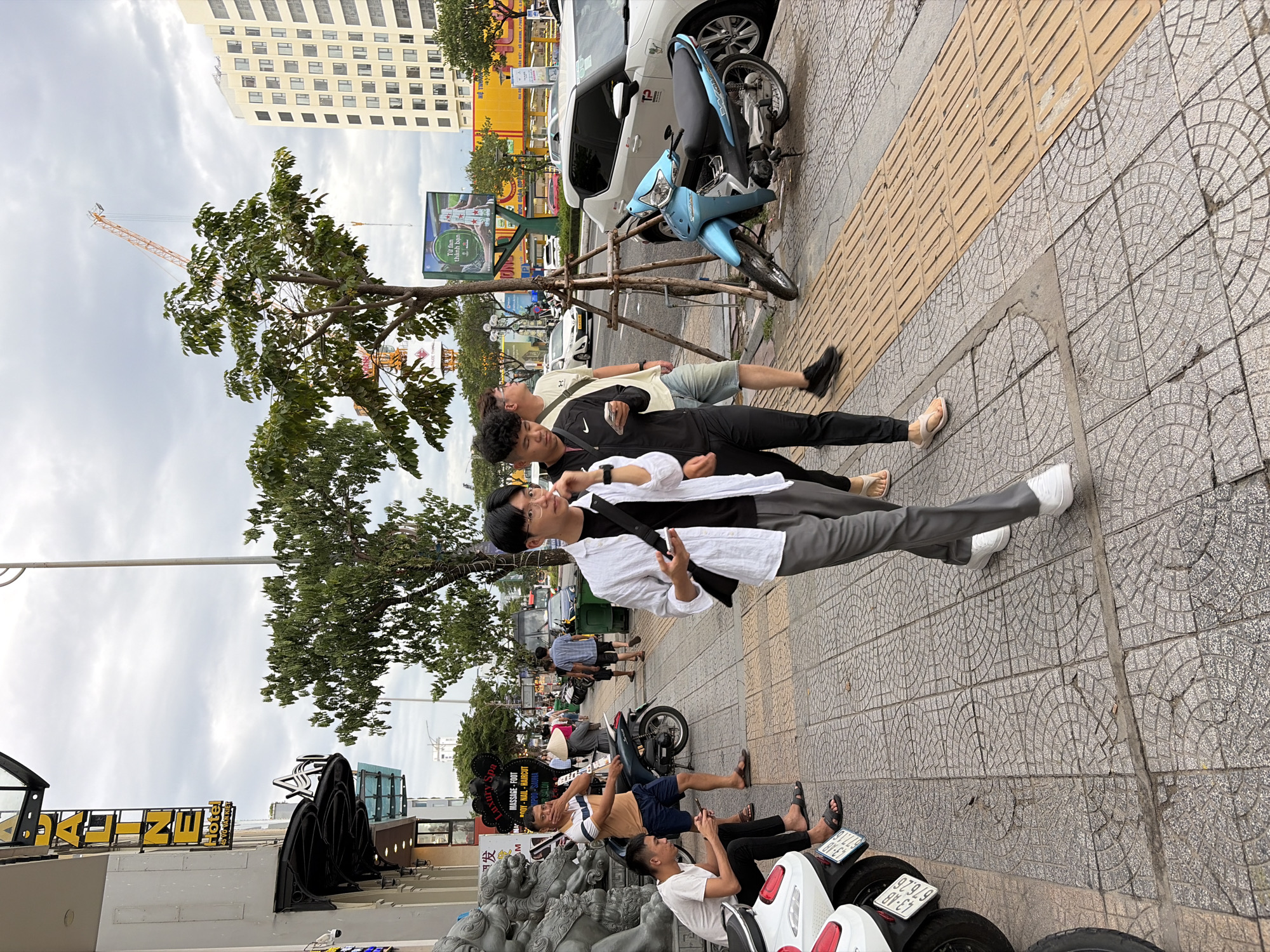 This is a photograph taken on a street in Da Nang, Vietnam. The scene depicts a sidewalk with several people, vehicles, and buildings. In the foreground, a young man in a white open-neck shirt and gray pants is walking while looking at his phone. To his left, another young man in a black jacket and black pants is also looking at his phone. Further to the left, two men are sitting on a motorcycle. One is wearing a blue and white striped shirt and blue shorts, and the other is wearing a white t-shirt and black pants. A teal motorcycle is parked on the sidewalk to the right of the foreground man. In the background, cars, trucks, and a bus are visible on the street. Buildings line the street, including a tall, modern building on the right and a building with a yellow sign that reads "JALINE Hotel" on the left. Trees and a construction crane are also present in the background. The sky is overcast.