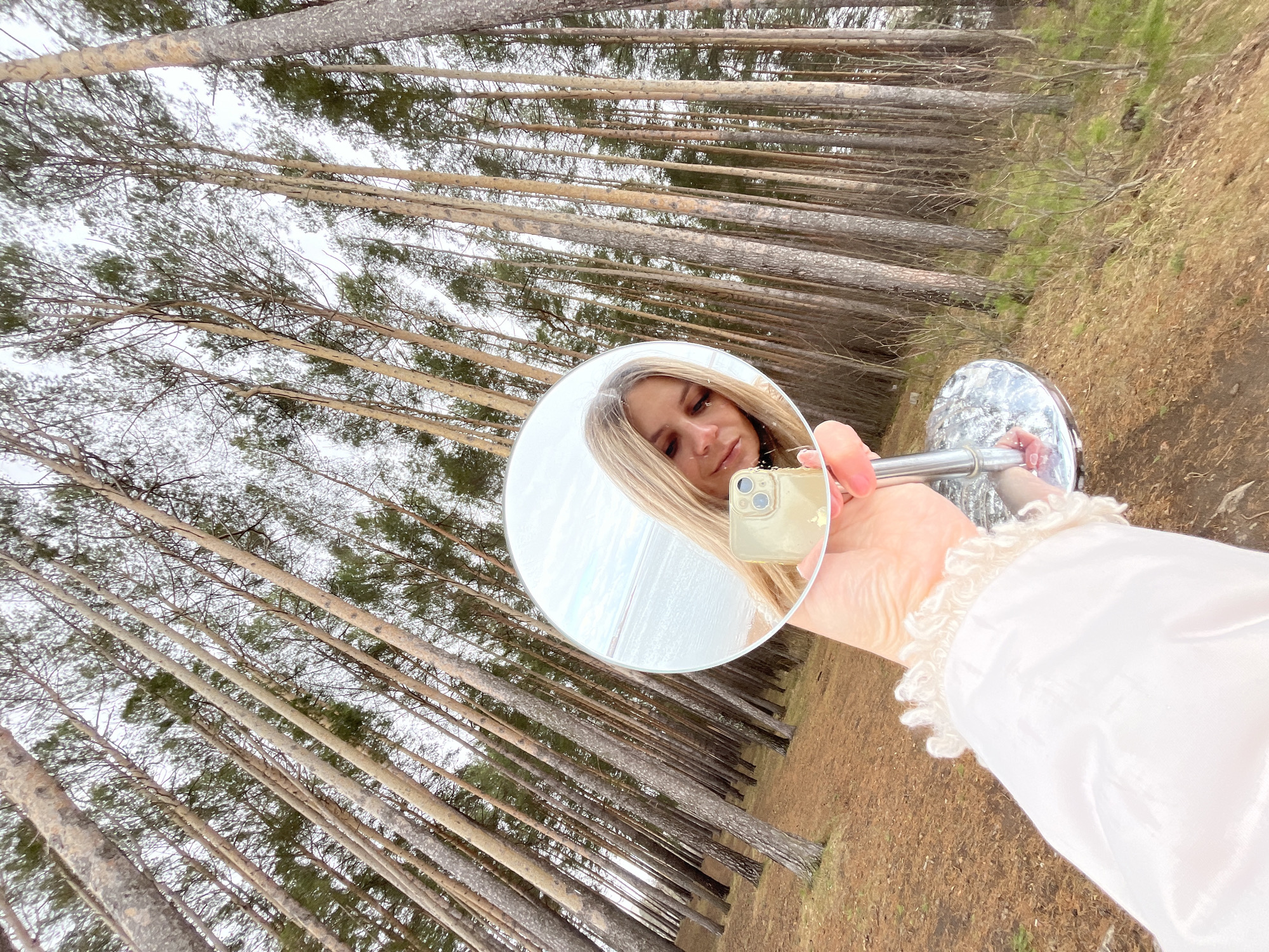 The image shows a woman holding a round mirror, reflecting her face and a distant beach, in a forest of tall, thin trees with brown trunks and green tops. The ground is covered with brown pine needles. The person's face is visible in the mirror, and she holds a gold-colored smartphone. The person wears a white blouse with lace trim on the sleeve. The location is Chaikovsky, Russia, based on the environment, suggesting a wooded area near a body of water.