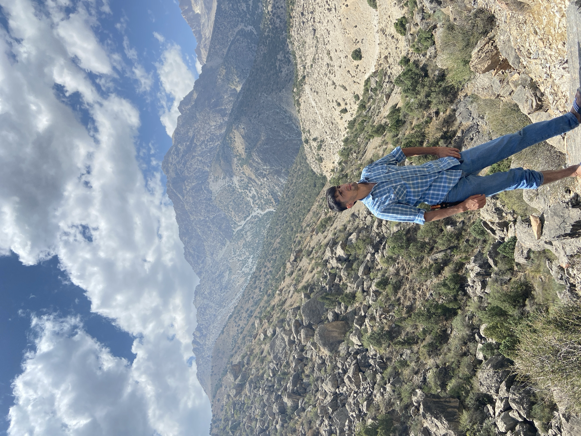 A young man in a blue plaid shirt and blue jeans stands on a rocky slope, looking up and to the right. Behind him, a vast mountainous landscape unfolds under a partly cloudy sky. The terrain is characterized by scattered rocks, sparse vegetation including small trees and bushes, and rugged, eroded mountain faces in the distance. The scene is likely outdoors in a natural, mountainous environment.