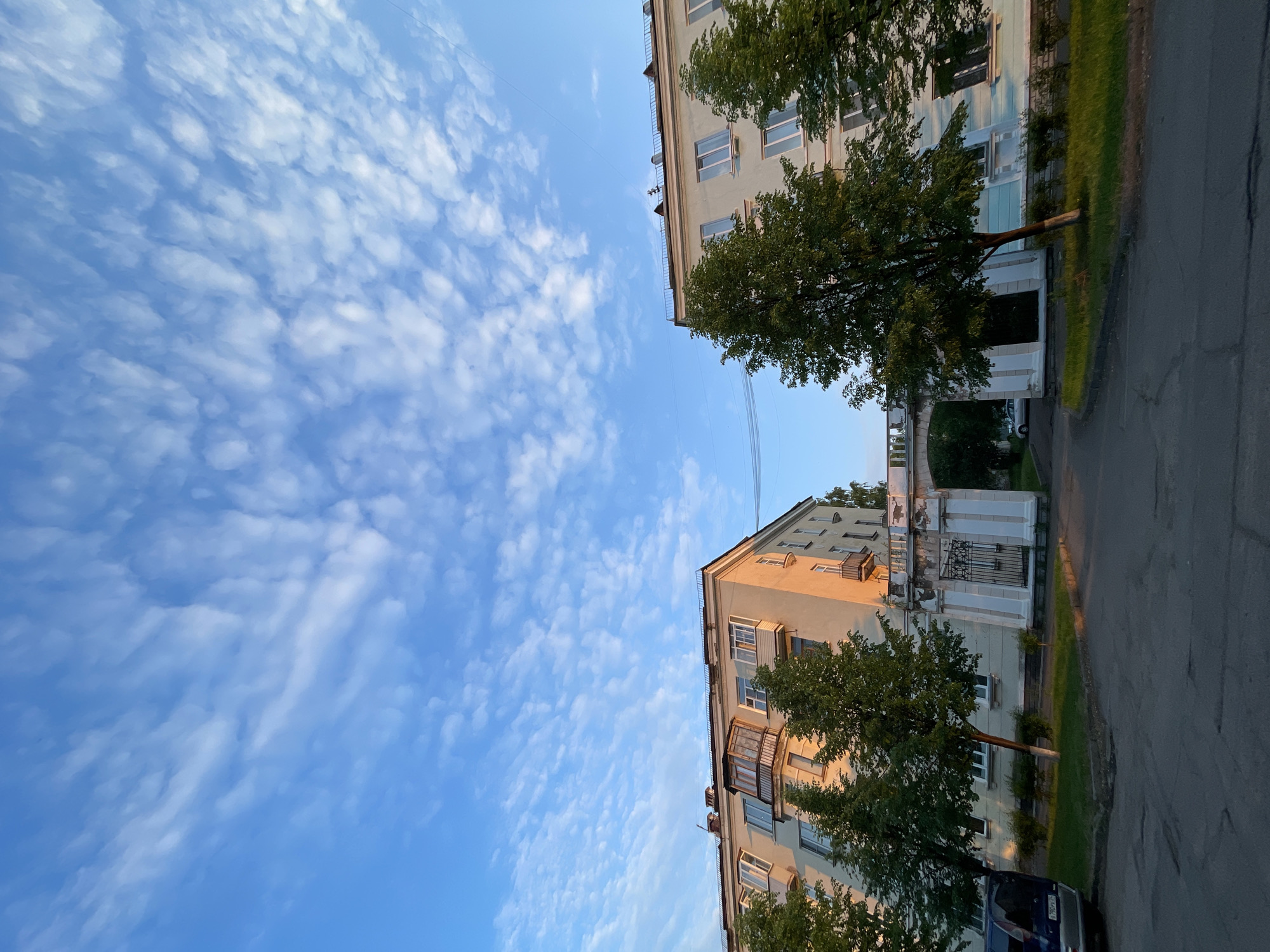 An outdoor, eye-level, full shot captures a pair of residential buildings in Zheleznogorsk, Russia, during the golden hour. The buildings, constructed in the Soviet-era style, face each other across a courtyard, connected by an arched gateway. The building on the left is a pale yellow, while the building on the right is a light blue. Both buildings have multiple stories, with balconies and windows visible. Trees with green foliage are planted in front of both structures, casting shadows on the asphalt courtyard. The sky above is a vibrant blue, filled with scattered white clouds that catch the warm light of the setting sun. The scene is quiet and peaceful, with no people or vehicles immediately visible in the frame.