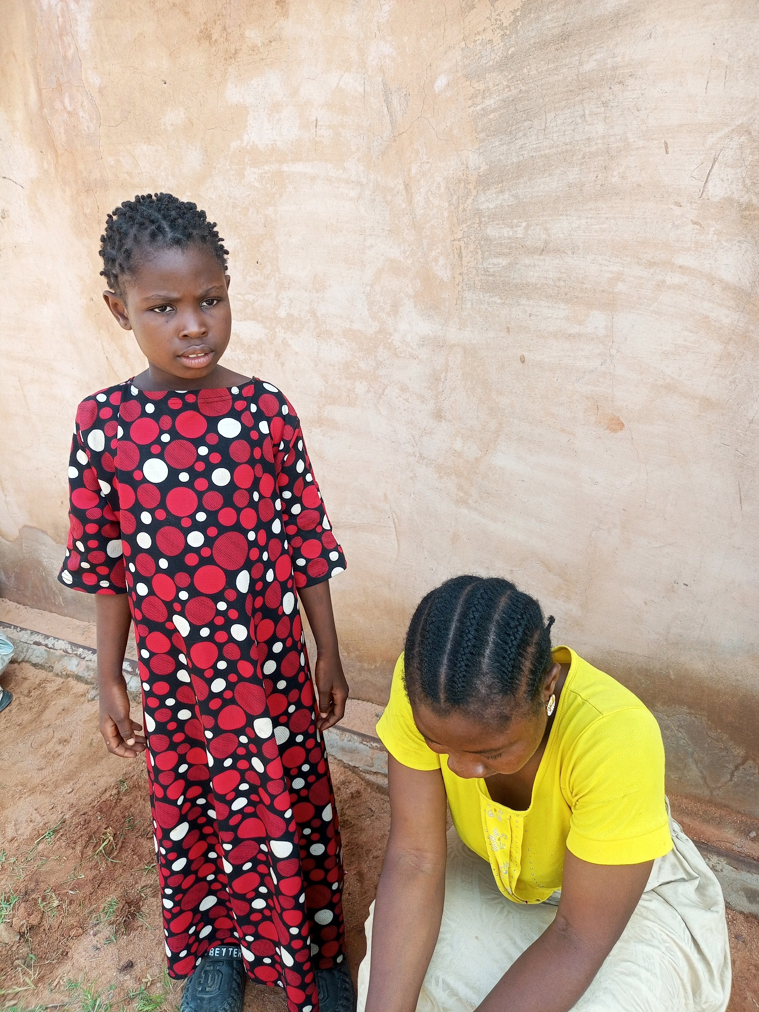 In Tempure, Nigeria, a young girl stands looking directly at the camera, her face serious. She wears a long, black dress with a pattern of red and white circles. Her hair is braided in many small plaits. To her right, a woman kneels, looking down. She is wearing a bright yellow t-shirt and a light-colored skirt. The two are positioned in front of a textured, light brown wall, with sand and dirt on the ground at their feet.