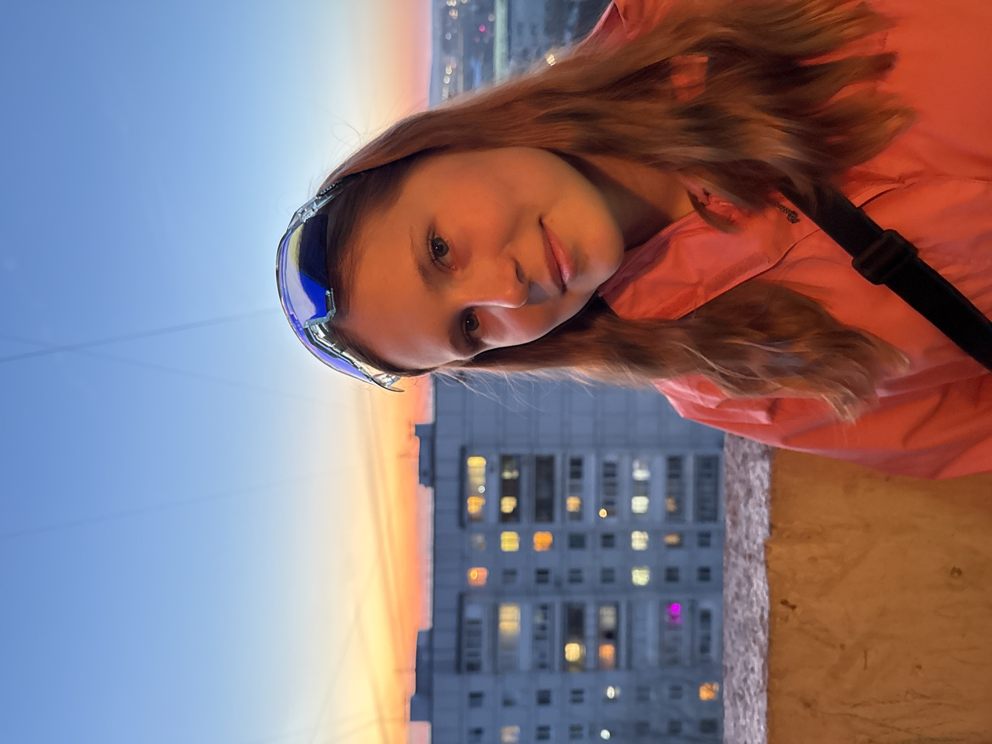The image is a selfie of a young woman, likely a teenager, with light brown hair and fair skin, wearing a light pink jacket and a pair of blue goggles pushed up on her forehead. The background features a high-rise building with numerous windows, some illuminated, and a sunset sky. The buildings suggest that the location is in an urban area, but the exact city cannot be conclusively determined. The woman is positioned on what appears to be a balcony or ledge.