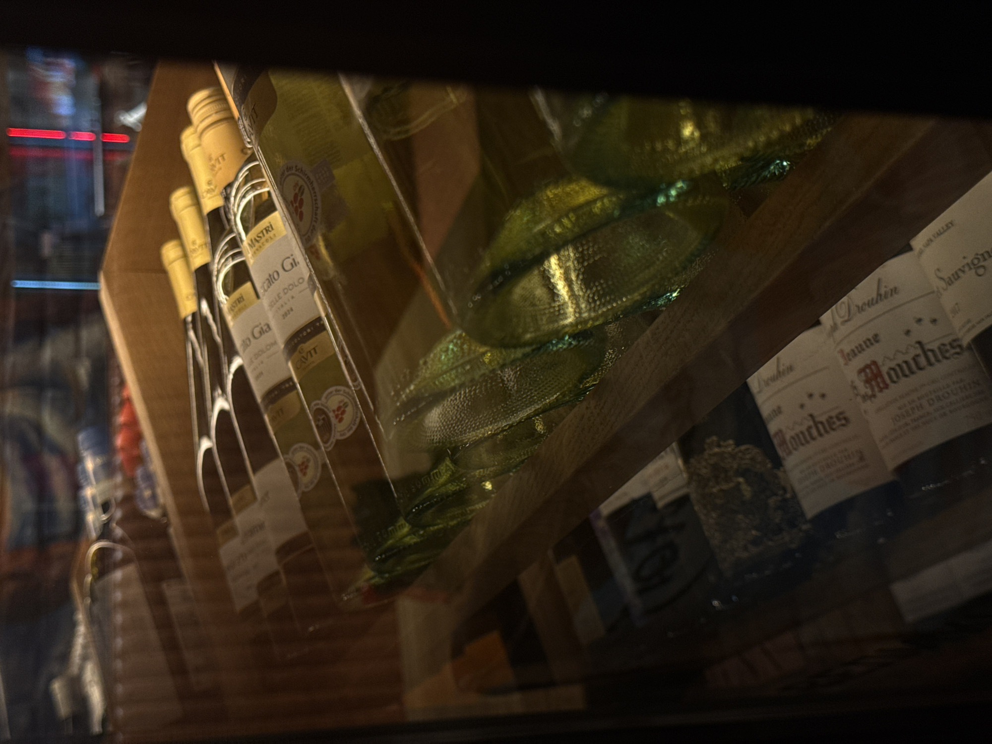 The image provides a medium close-up, slightly angled view of multiple bottles of wine stored horizontally on wooden shelves within a display cabinet or cooler, viewed through a reflective glass front. The scene is dimly lit, with internal display lighting and prominent reflections from the surrounding environment.
On the upper shelf, approximately six white wine bottles are discernible, each featuring a yellow foil cap. Labels on these bottles are partially visible, with clear identification of "Moscato Gia," "DOLOMITI," and the vintage "2021" on some, and "MASTRI" on another, indicating Italian origin. The wine within these bottles appears light yellowish-green.
On the lower shelves, additional wine bottles are visible. Some bottles are made of green glass, others of lighter glass. Labels are more difficult to read due to reflections and lower lighting, but fragments show "Mouches," "JOSEPH DROUHIN," and "Sauvignon," suggesting French wines, including a known Burgundy producer.
The glass front of the display cabinet shows significant reflections. In the upper background, highly blurred areas of red vertical streaks (possibly neon or LED signs) and other diffuse blue and white lights are reflected, suggesting an urban or indoor setting outside the immediate display. The lower portion of the image is further obscured by reflections, creating iridescent green and yellow hues from the bottles and diffuse light patterns.
The shelving system consists of light-colored wooden slats and vertical supports. No individuals are visible in the image. The setting is consistent with a commercial establishment, such as a wine shop, restaurant, or supermarket, in the city of Izhevsk, Russia, although no specific visual cues within the image directly identify Izhevsk.