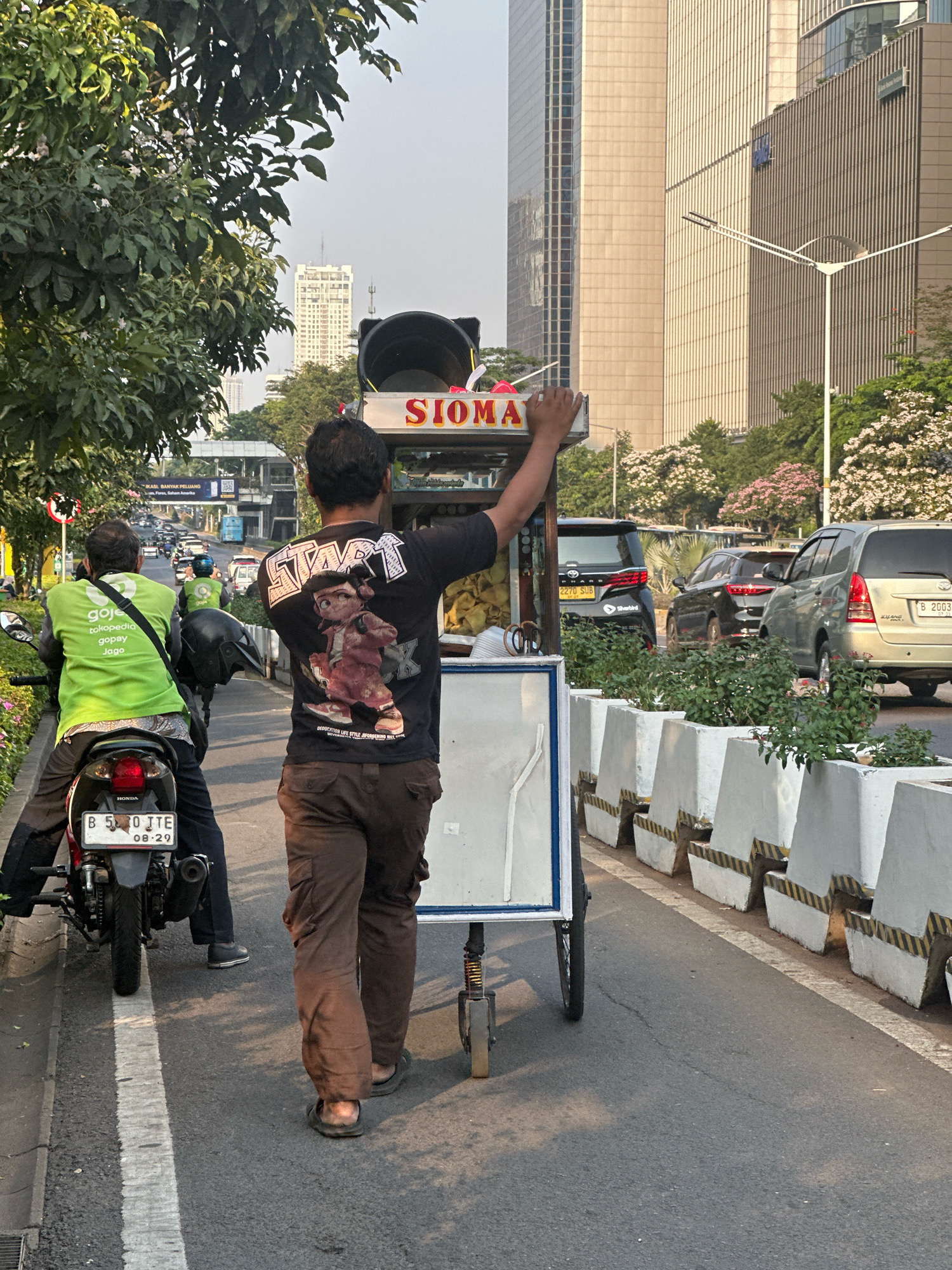 A street vendor pushes a siomay cart on a busy street in South Jakarta, Indonesia. The vendor, wearing a black t-shirt with a cartoon character graphic and brown cargo pants, is walking away from the camera. The cart has a sign that reads "SIOMAY" in red lettering. To the left, a person wearing a bright green Gojek uniform is seated on a motorcycle. In the background, modern buildings rise above trees, and traffic can be seen on the road. The scene appears to be set during the daytime, with sunlight casting shadows on the street. The street is lined with planters and concrete barriers.