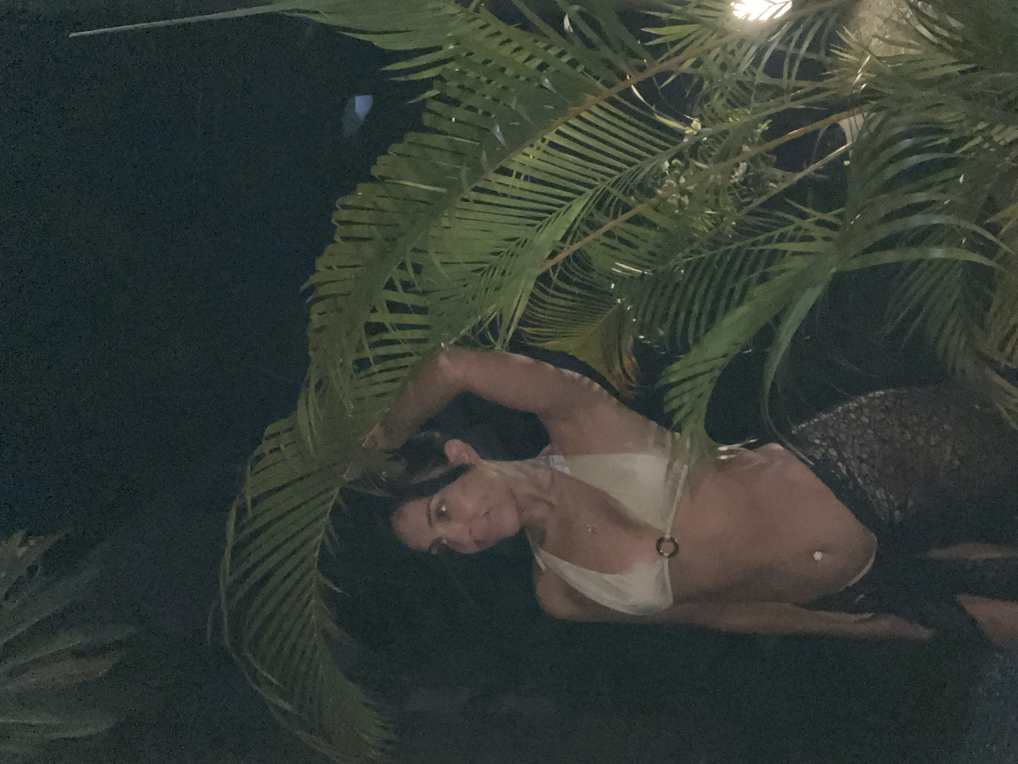 A woman is reclined outdoors in Kuta Selatan, Indonesia. She is wearing a white top, with dark colored bottoms. Palm fronds partially obscure her, and the scene is dimly lit.