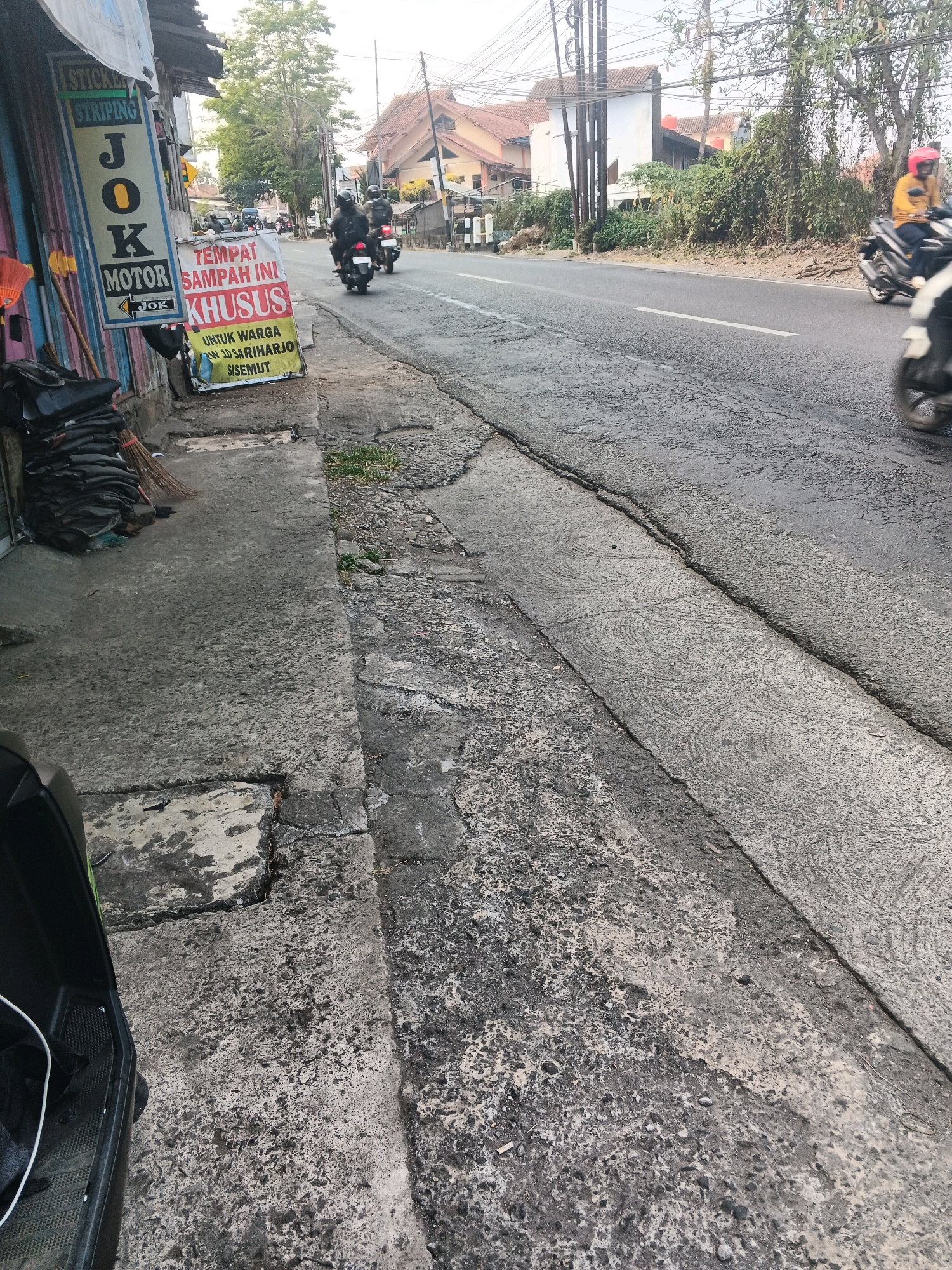 This image depicts a street scene in Ungaran, Indonesia, likely near a shop. On the left side, a sign for "JOK MOTOR" and "STICKER STRIPING" is visible, along with stacked motorcycle seats and a broom. A banner with Indonesian text, "TEMPAT SAMPAH INI KHUSUS UNTUK WARGA 3D SARIHARJO DISEMUT," which translates to "This trash can is specifically for residents of 3D Sariharjo DiseMut," is also displayed. Several motorcycles are present: some parked on the sidewalk, and others in motion on the road. One motorcycle in the foreground on the left appears to be partially visible, with a black body and a white cable. The road is paved and shows signs of wear, with cracks and an uneven surface. The environment suggests a typical urban setting with buildings and vegetation visible in the background. The lighting indicates it is daytime.