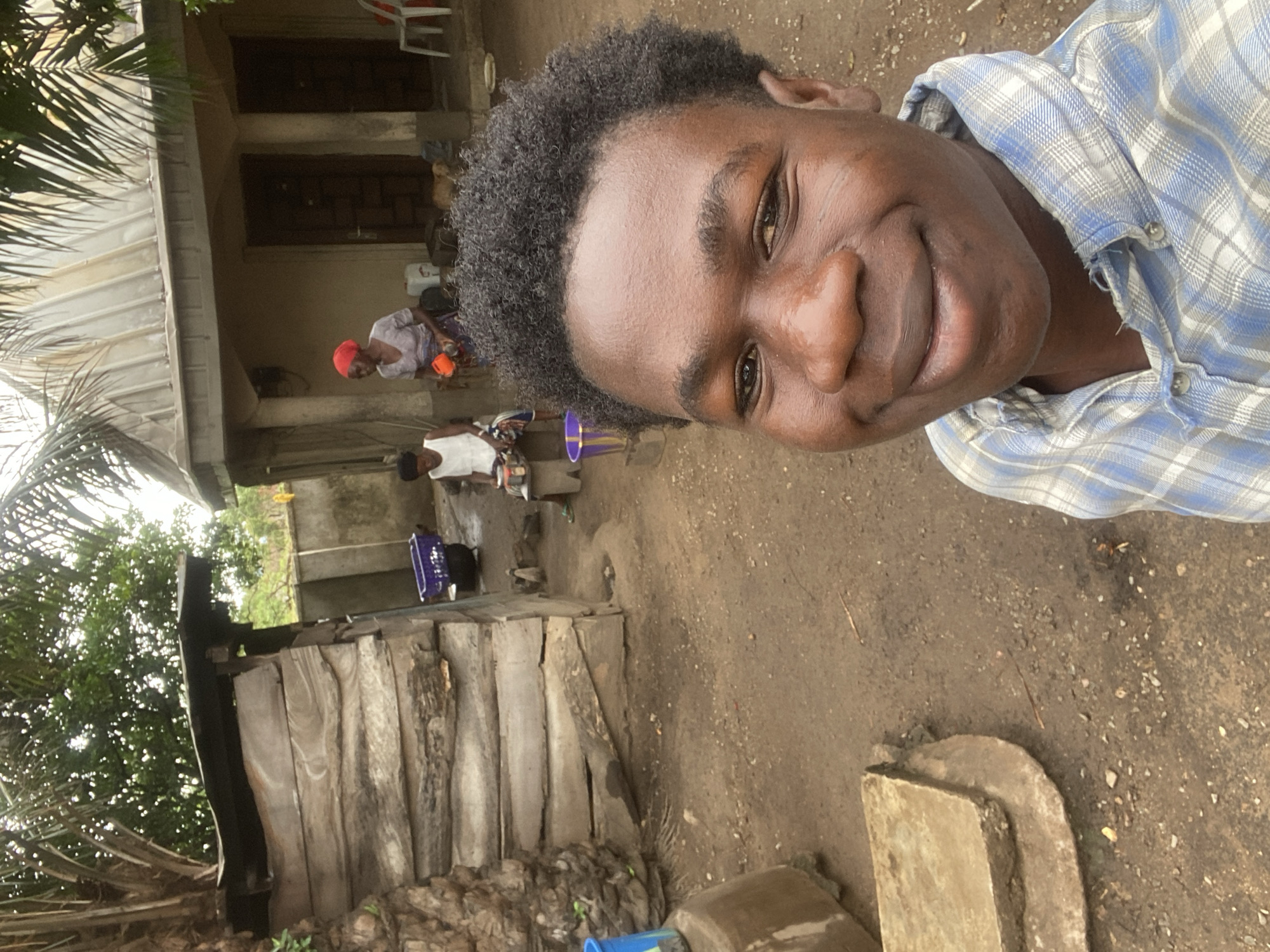 The image depicts a close-up of a young Black man with short, dark curly hair, smiling at the camera. He is wearing a blue and white plaid shirt. The background includes a dirt ground, and a partially visible wooden structure, possibly part of a dwelling, with a set of uneven wooden steps. Further back, two women can be seen near a house, one wearing a red headscarf. Based on the information provided, this scene is in Mararaba, Nigeria. The environment appears to be a rural or less developed area.