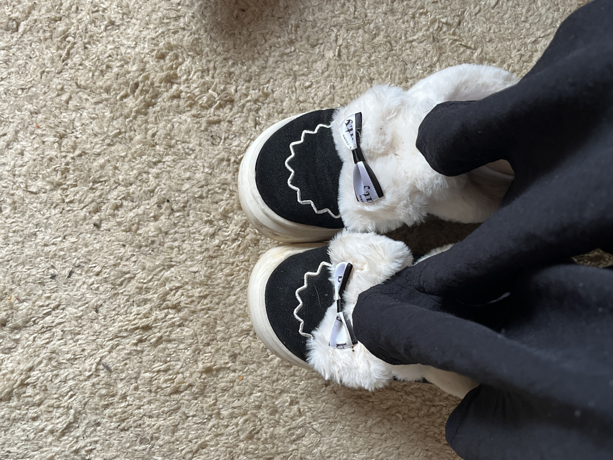 The image shows a pair of slipper shoes on a beige carpet. The slippers are black and white, with white faux fur trim and a small white ribbon on each. A leg covered in black fabric is also visible, seemingly wearing the slippers. The location is unknown.