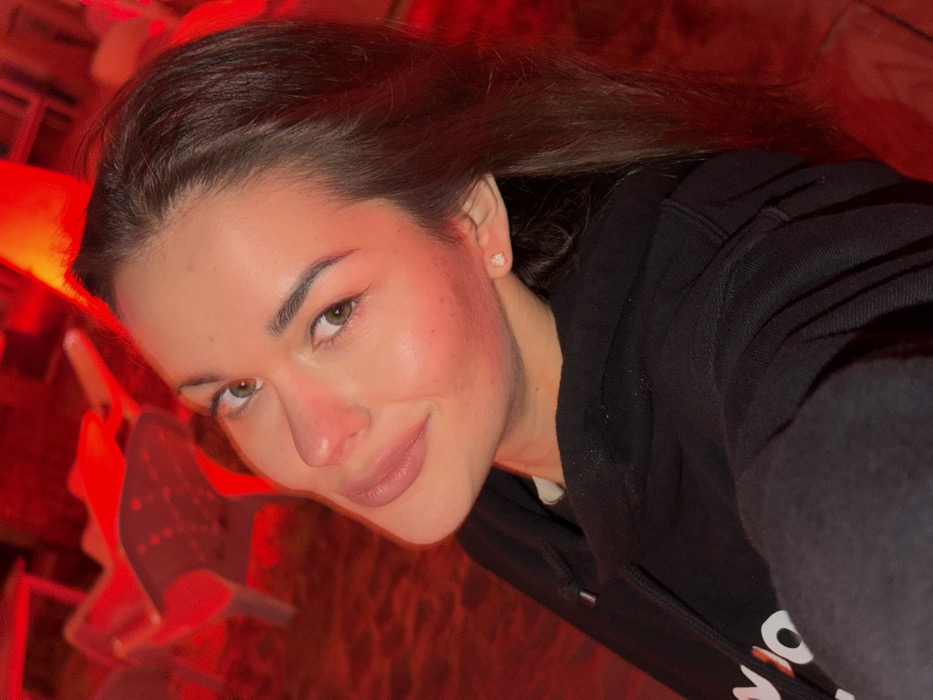 A female subject is depicted in a close-up portrait, oriented slightly to the left and looking directly at the camera, within an establishment in an urban setting consistent with Tel-Aviv, Israel. The environment is dominated by strong red lighting, casting a deep crimson hue across the scene.
The subject is a young adult female with light skin, dark brown hair, dark eyebrows, and light brown or hazel eyes. She presents a slight, closed-lip smile. Her right earlobe contains a small, round, light-colored stud earring. She is wearing a black hooded sweatshirt, beneath which a white inner collar is visible. The sweatshirt fabric, particularly on the right shoulder, exhibits small, light-colored particles.
The background is out of focus and heavily saturated with red light. Visible objects include indistinct, light-colored chairs or furniture pieces, some appearing to be stacked or inverted, consistent with a casual commercial space such as a bar, club, or restaurant patio. The floor or ground surface in the background appears textured. No other individuals are present or engaged in interaction with the primary subject within the frame.