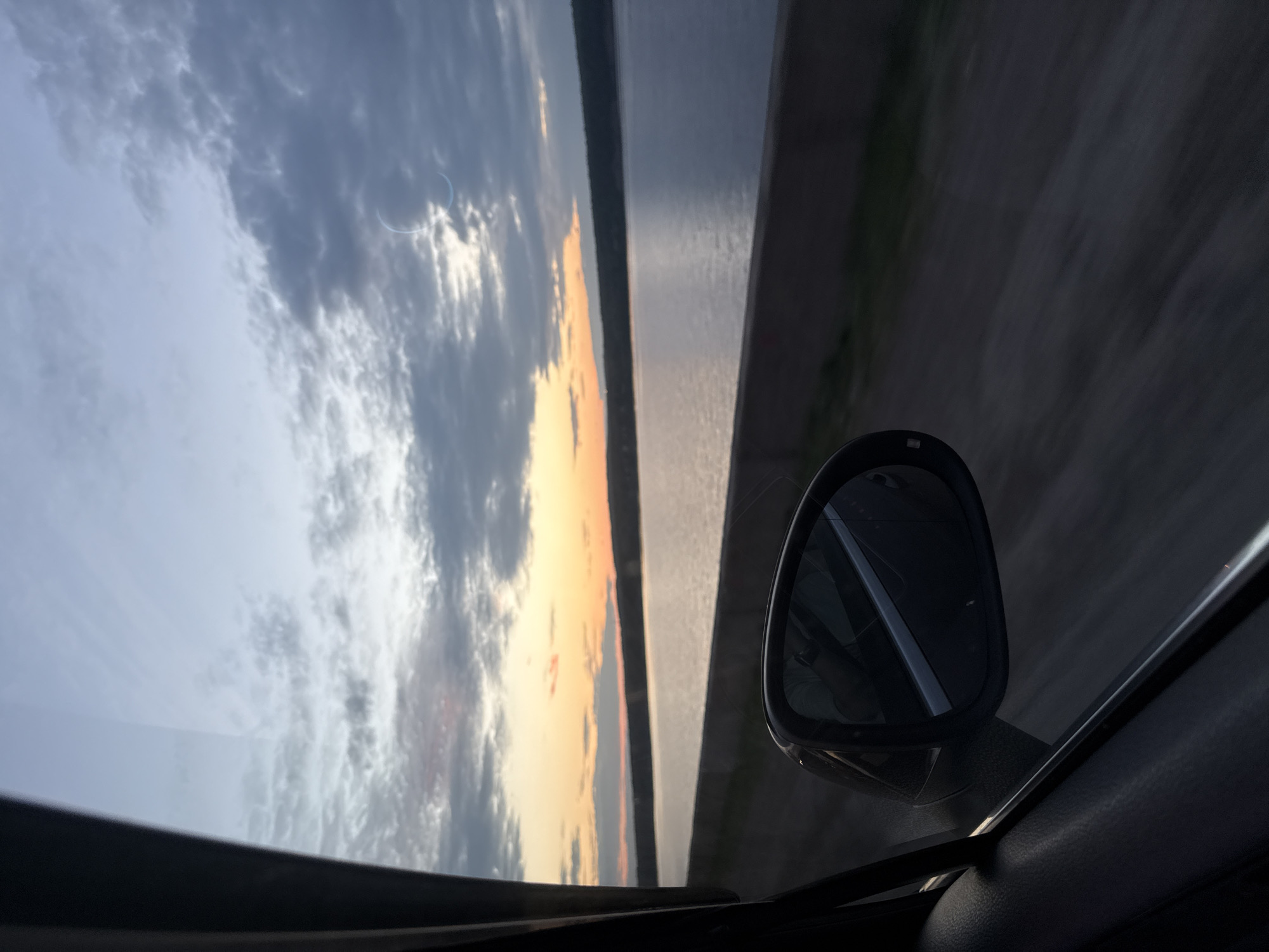 A view from inside a moving vehicle shows a body of water with a treeline on the opposite shore under a cloudy sky at dusk. A car's side mirror is visible in the foreground, reflecting the interior of the car. The sky displays shades of orange and grey, with the sun low on the horizon. The water is calm and reflects the colors of the sky. The image captures a scenic view of a natural landscape, possibly along the Kama River near Chaikovsky, Russia.