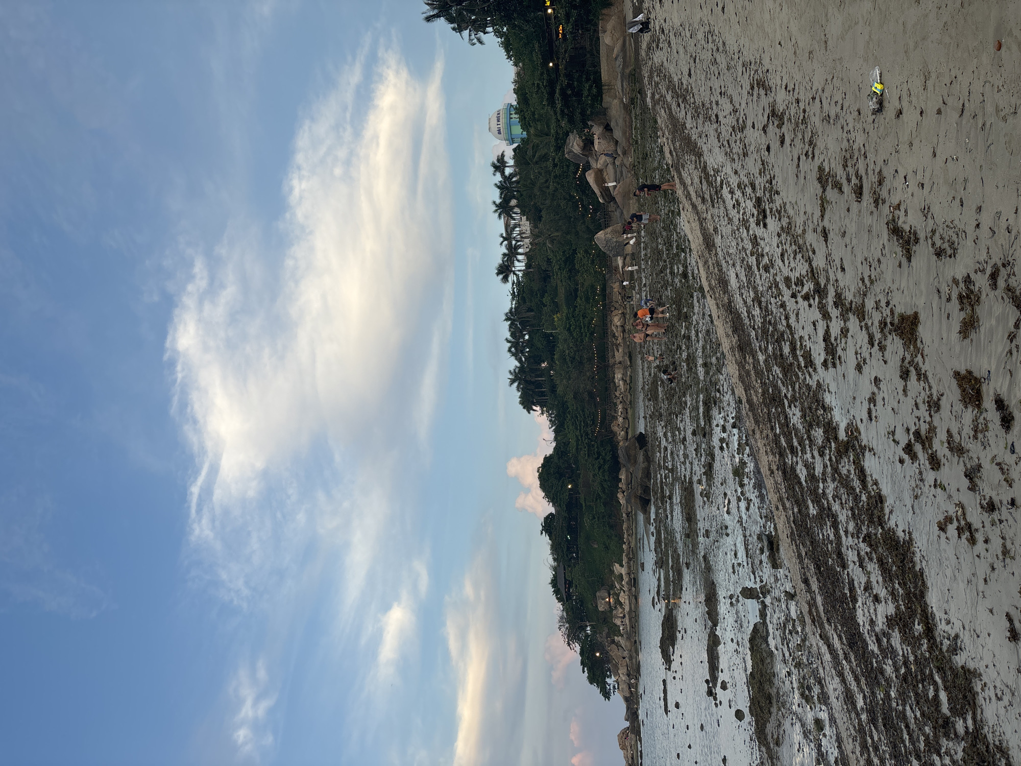 The image shows a coastline in Bac Nha Trang Ward, Vietnam. The sky is partly cloudy with blue sky visible. The beach is littered with seaweed and debris. Several people are present on the beach, some appearing to be gathering items or interacting with the environment. The coastline is lined with trees and vegetation, and a building with the words "ĐẠI THIÊN LỘC" is visible in the distance. The overall impression is a coastal scene at dusk or dawn.