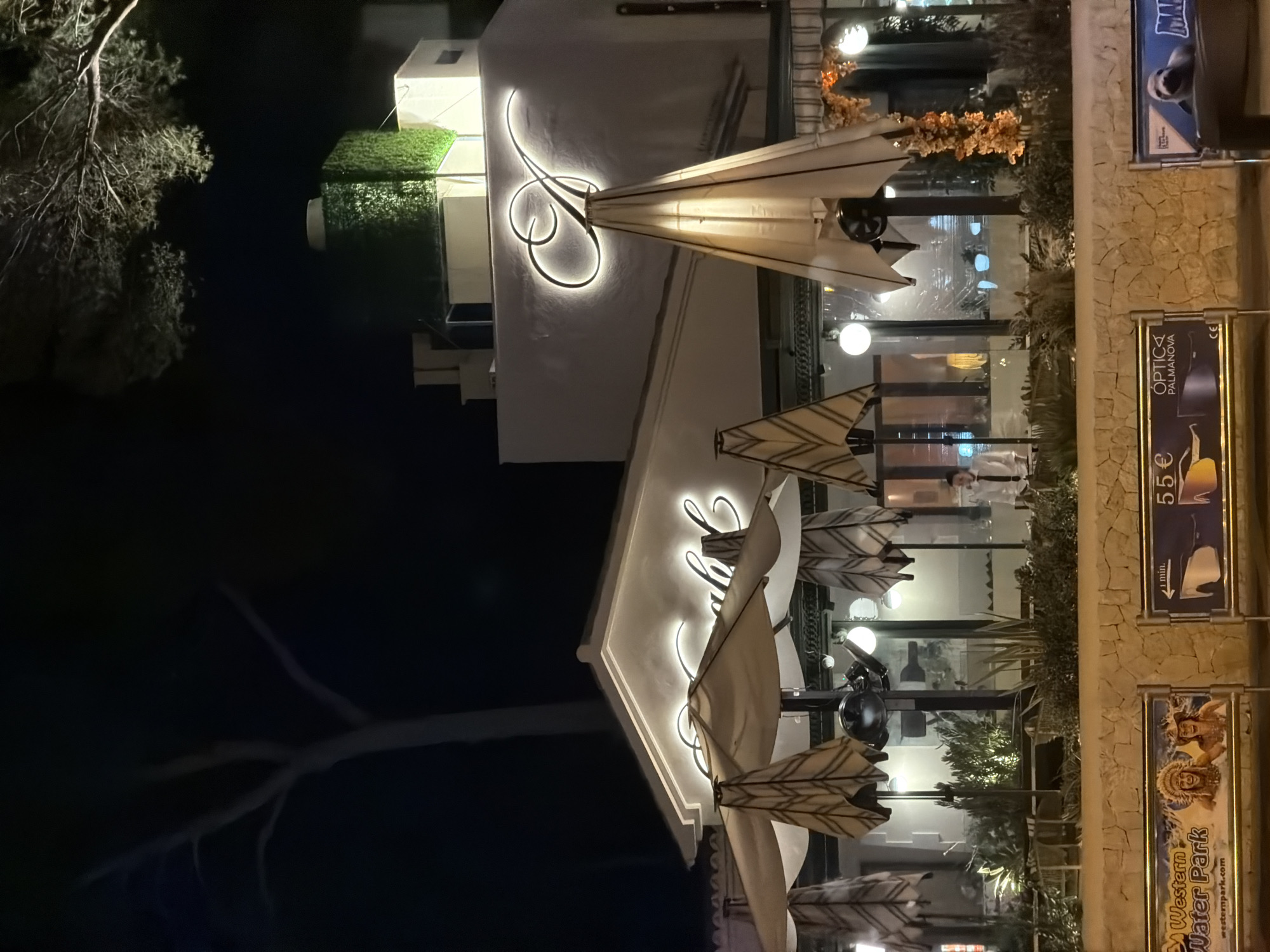 The image shows the exterior of a restaurant at night in Palmanova, Spain. The restaurant has a white facade with illuminated script signage that reads "A" on the upper level and what appears to be "Sailor" on the lower level. There are large umbrellas and outdoor seating areas visible in front of the restaurant. A person in a white shirt and dark tie is standing inside the restaurant behind a counter. In the foreground, there is a low stone wall with promotional signs. One sign advertises "Western Water Park" with images of two smiling people. Another sign shows "Optica Palmanova" and a price of "5€." A third sign features a seal and the word "MAR." The lighting suggests evening or nighttime.