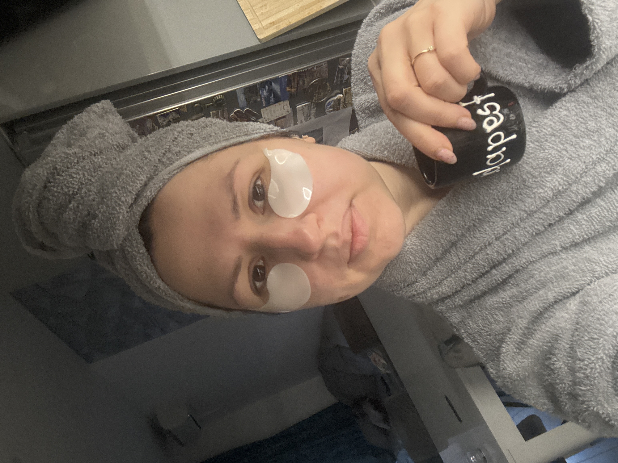 A woman in a gray bathrobe and towel headwrap is holding a black mug with white writing on it. She has white patches under her eyes. She is wearing a gold ring. The scene is indoors, with a metal panel behind her head that has magnets and miscellaneous papers stuck to it. There is a cutting board above the metal panel. In the background, there is a room with blue curtains and a white wall. The location is not identifiable as Sofia, Bulgaria.
