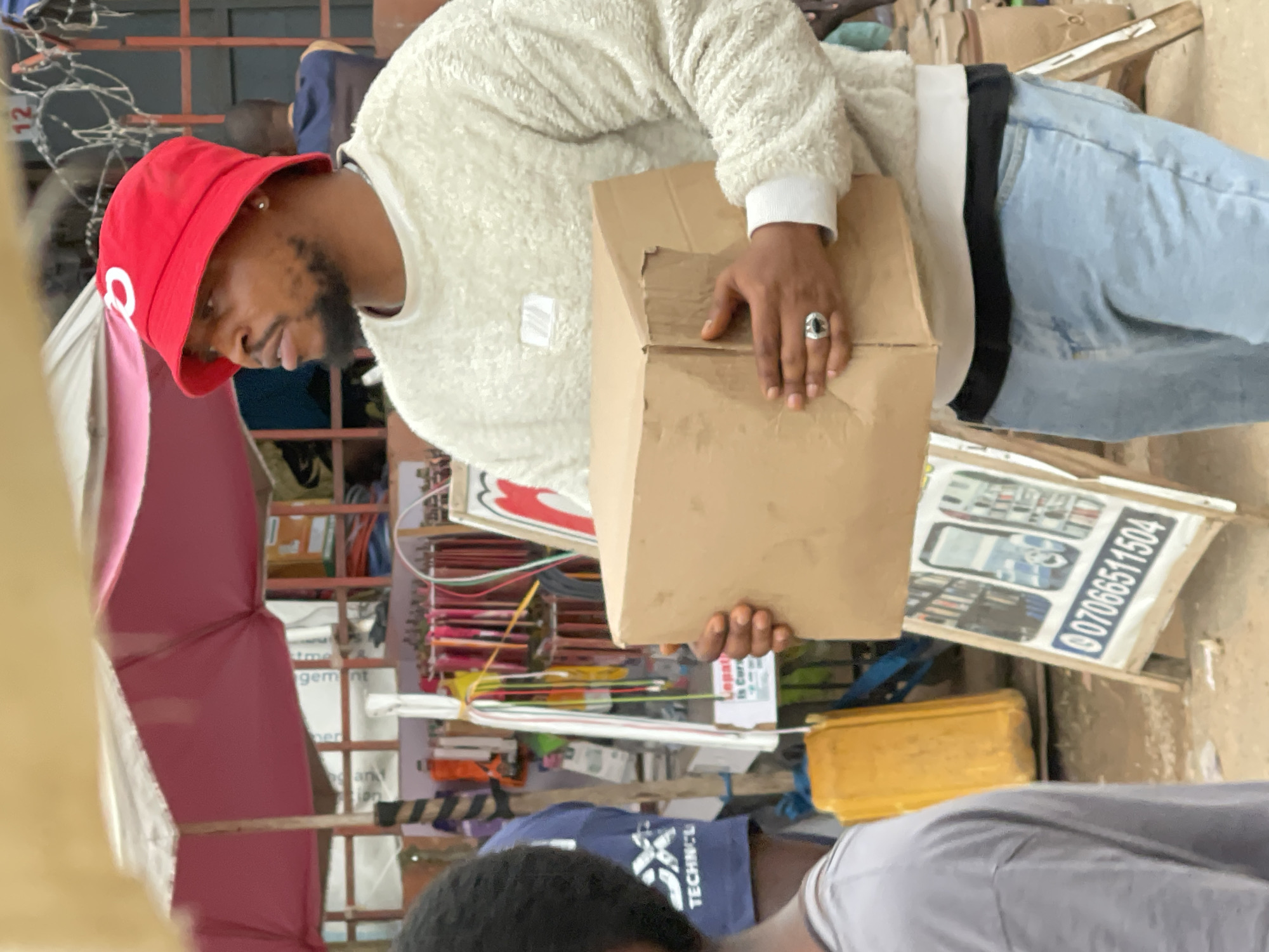 A man wearing a red bucket hat and a cream-colored fuzzy sweater is holding a cardboard box in what appears to be an outdoor market stall in Abuja, Nigeria. He is also wearing light blue jeans and a silver ring with a black stone on his left hand. In the background, there are market stalls with various items for sale, including clothing and what looks like a large yellow container. A portion of a pink umbrella is visible overhead. A sign with a phone number and images of phones is propped up on a wooden easel.