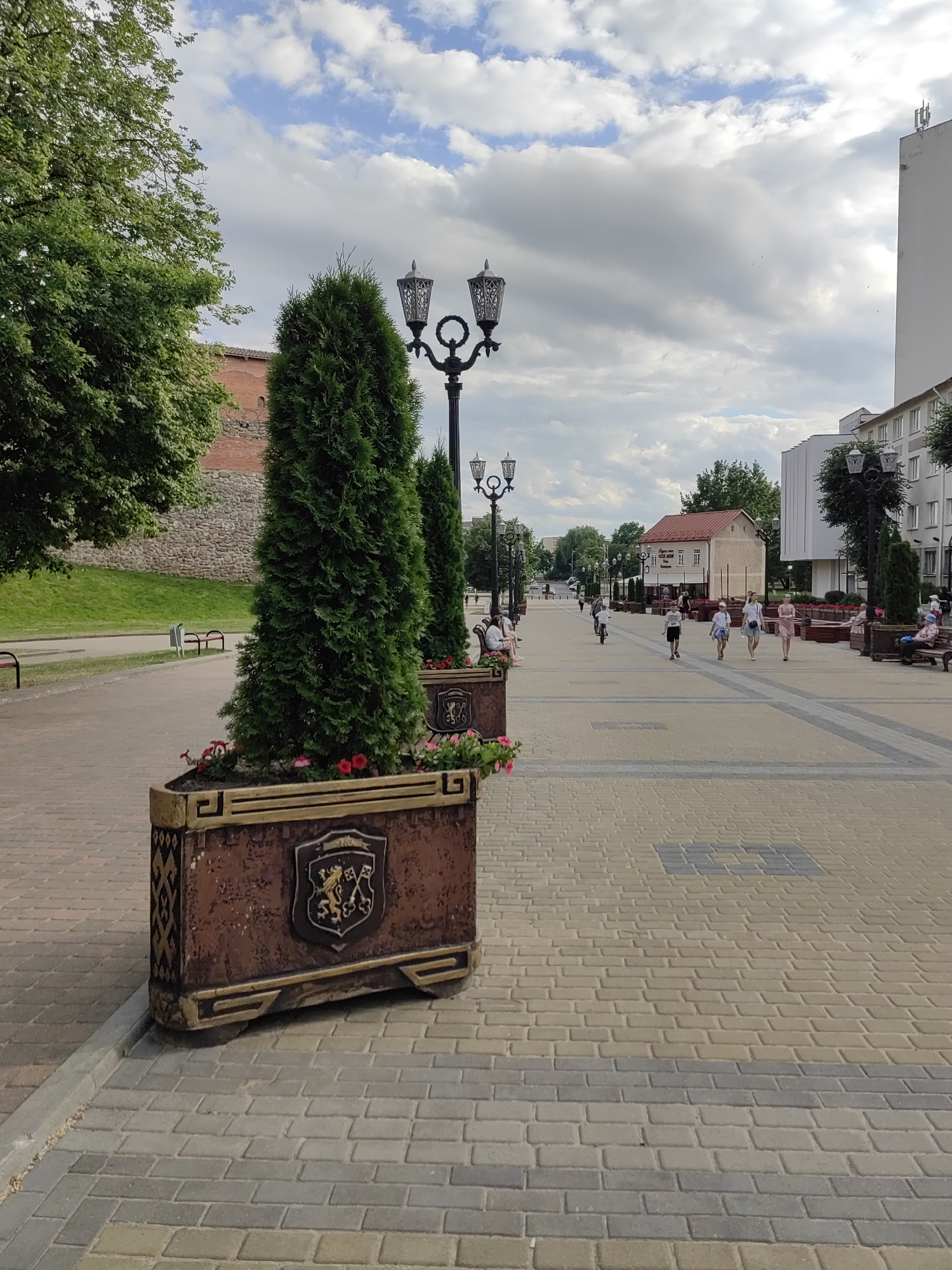 This media file captures an outdoor scene along a paved walkway lined with trees and decorative planters. The walkway is paved with light-colored rectangular bricks, interspersed with strips of darker grey bricks. Several lampposts with ornate designs are visible along the walkway.
In the foreground, a large, rectangular planter with a dark brown, weathered finish and gold trim contains a tall, green coniferous tree and some red flowers. A crest with a lion and keys is visible on the side of the planter. Behind this, another similar planter and tree are partially visible.
Further down the walkway, several people are strolling. A family of four, including two adults and two children, walk towards the viewer. A child rides a bicycle further ahead. Other individuals are seated on benches along the sides of the walkway.
In the background, a red brick wall with a grassy slope behind it is visible on the left. On the right, modern buildings and a smaller building with a red roof stand. The sky is partly cloudy with patches of blue and grey clouds, suggesting it might be daytime. There is no visible text in the image. The overall impression is a pleasant, leisurely atmosphere in a town or city setting.