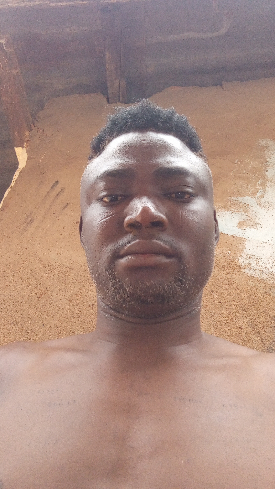 A medium shot of a bare-chested Black male with a close-cut hairstyle, facing the camera. He has facial hair and a scar on his left cheek. The background is a textured beige wall with some damaged areas and exposed wooden beams. The image appears to have been taken at eye level. The location is Sabon Tasha, Nigeria.