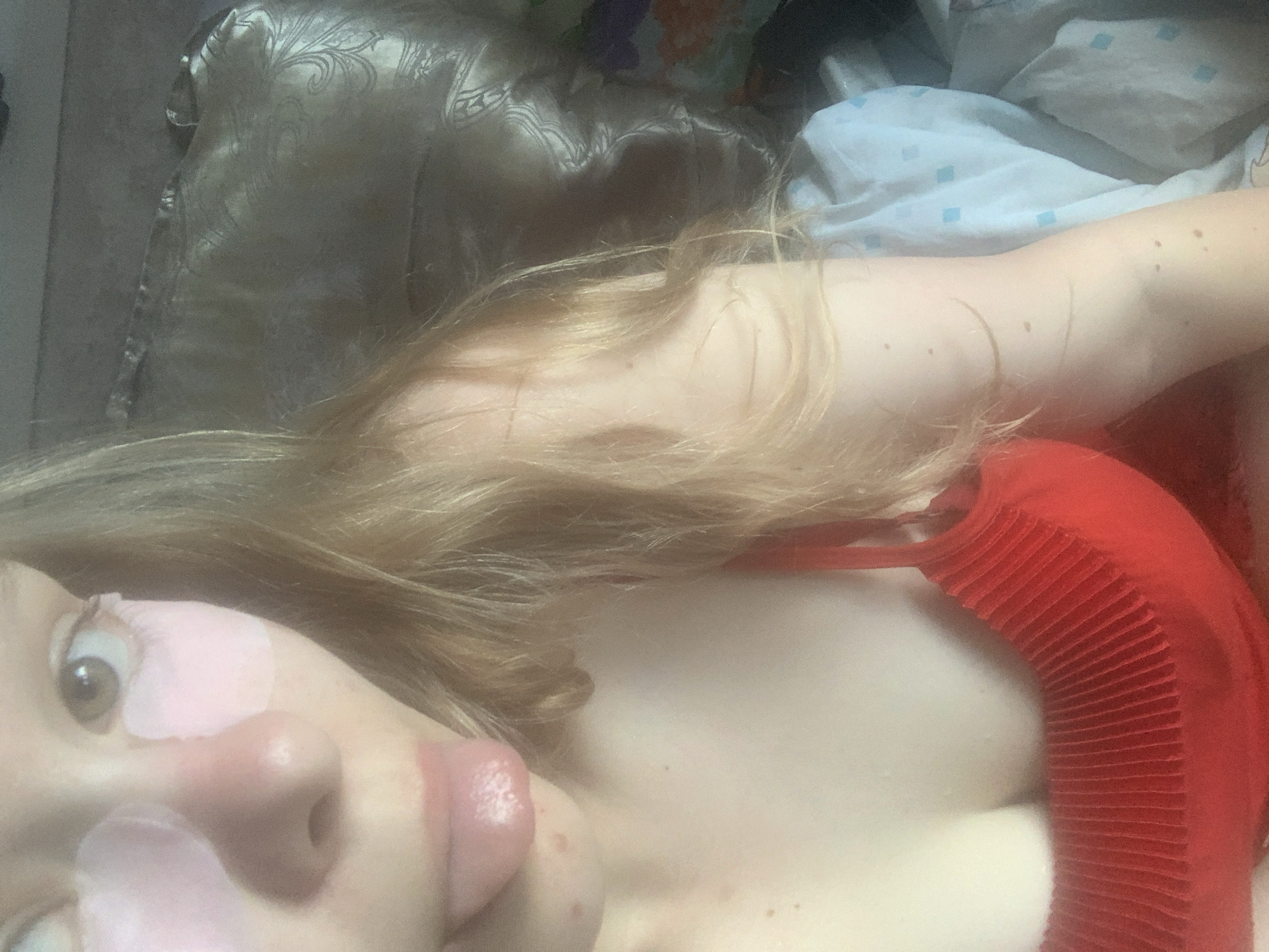 A close-up, indoor, medium shot shows a young blonde woman with pink under-eye patches. She is wearing a red, ribbed halter top. Her face is angled towards the camera, and her tongue is slightly extended from her mouth. The background includes a textured, possibly leather couch and a patterned blanket or sheet. The lighting is soft and somewhat diffused. No specific location in the city of Unknown, Russia can be determined from the image.