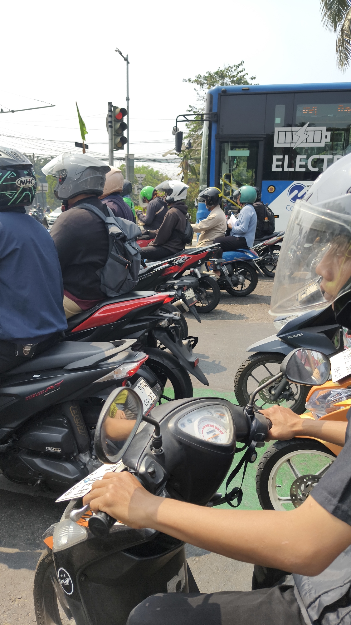 The image depicts a traffic scene, likely a traffic light stop, in South Jakarta, Indonesia. A blue electric bus with "ELECTRI" visible on its side is prominent in the background, alongside a traffic light showing red. The foreground is dominated by several motorcycles and their riders, many wearing helmets. One rider in the immediate foreground is partially visible, their hands on the handlebars of a black scooter, with a clear visor on their helmet. Another rider is on a red and black motorcycle, and further back, other motorcycles and riders are visible, including one on a blue scooter. The environment suggests an urban setting with roads, traffic infrastructure, and vegetation. The time of day appears to be daytime, with bright sunlight casting shadows.