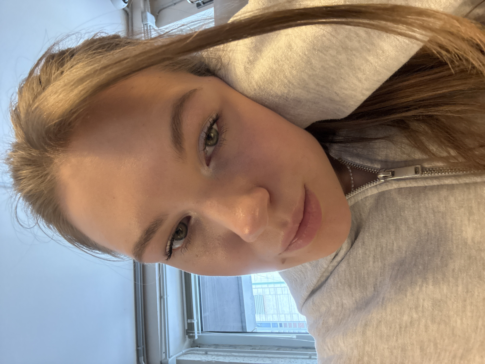 A close-up, angled selfie shows a young woman with light brown hair and fair skin wearing a gray hooded sweatshirt. She has green eyes and a neutral expression. A window is visible in the background, showing a generic urban scene with buildings. There is no information in the image to identify a specific location in Belgrade, Serbia.