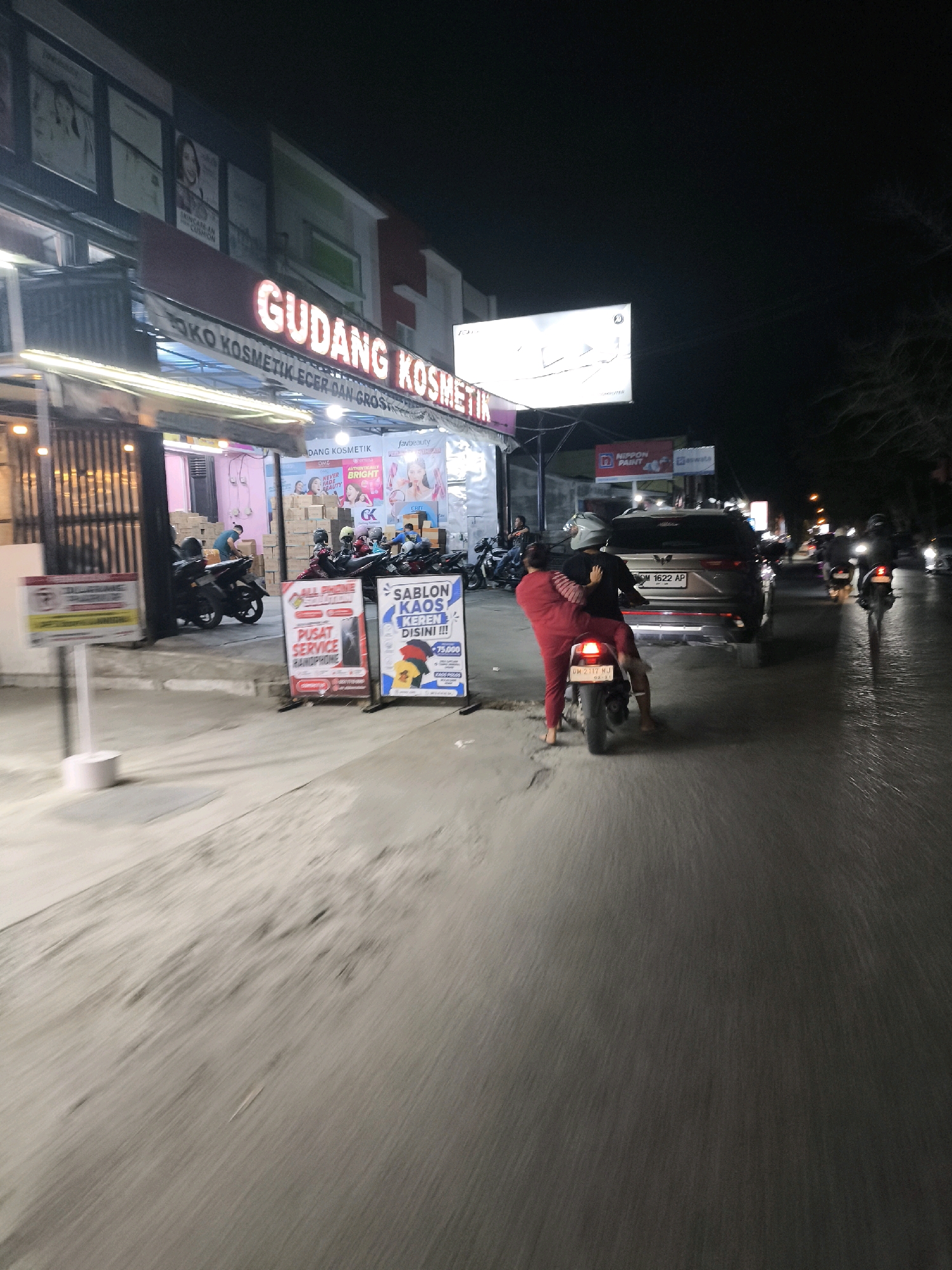 The image captures a night scene on a street in Gorontalo, Indonesia. A store identified as "GUDANG KOSMETIK" (Cosmetics Warehouse) with illuminated signage is prominently displayed. In front of the store, several motorcycles are parked. On the street, a person wearing a red outfit is riding on the back of a motorcycle, with their arms around the driver. A gray SUV is also visible on the road. The street surface appears wet, suggesting recent rain or damp conditions. Signs for "ALL PHONE SERVICE" and "PUSAT SERVICE HANDPHONE" are positioned on the sidewalk. The overall ambiance is that of a bustling commercial area at night.