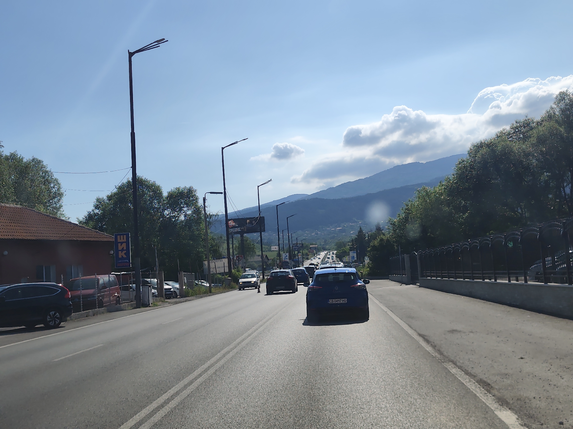 The image captures a view of a street in Dragichevo, Bulgaria, on a sunny day. The scene depicts a road with multiple lanes of traffic, bordered by trees and buildings on the left and a fence with decorative arches on the right. A blue car with license plate CB 0497 HO is in the foreground, driving on the right side of the road. Several other cars are visible on the road, moving in the same direction. In the background, mountains are visible under a sky with scattered clouds. On the left side of the road, a building with a red tiled roof and a sign with Cyrillic lettering is present, along with parked vehicles. Streetlights are positioned along the road. The overall impression is that of a suburban or semi-urban environment on a clear day.