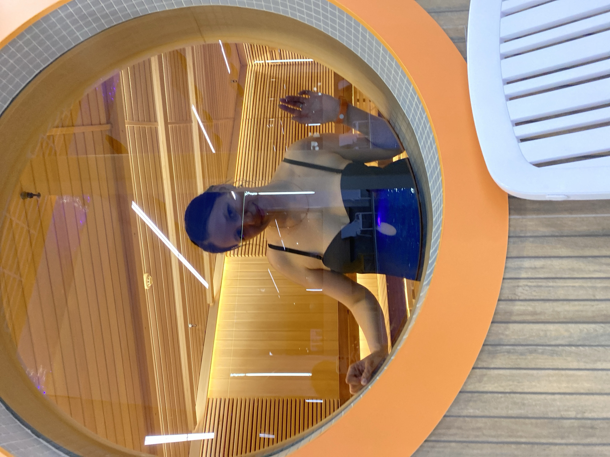 A reflection of a woman wearing a black swimsuit and blue bathing cap is visible in a circular glass enclosure. The woman's face is turned towards the viewer, and her hand is raised near the top of the enclosure. The enclosure is framed by an orange rim with a gray tile border, set against a backdrop of horizontal wooden planks. Visible within the enclosure is a modern architectural interior featuring vertical wooden slats and integrated lighting. To the right of the enclosure, a white, slotted object is visible. The location is Votkinsk, Russia.