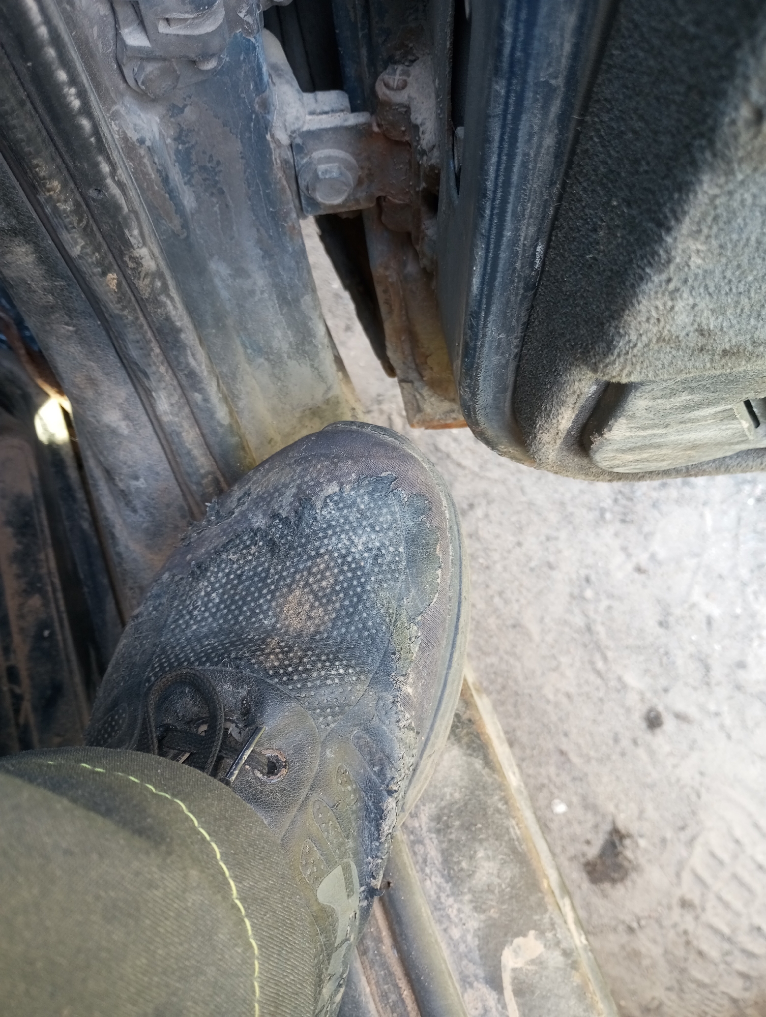 A close-up, slightly angled shot shows a person's foot wearing a dirty, dark-colored work boot. The boot's toe area is textured and shows signs of wear and tear. The boot is resting on a metal surface, possibly a vehicle's step or frame. To the left of the foot, a dark, ribbed rubber or plastic material is visible, likely part of the vehicle's interior or exterior trim. To the right of the foot, the ground appears to be a light-colored, dusty surface, possibly concrete or dirt. In the background, a dark metal structure with bolts and some rust is visible, suggesting a mechanical or industrial environment. The overall scene appears to be outdoors or in an industrial setting, possibly in Alabukhino, Russia, given the context.