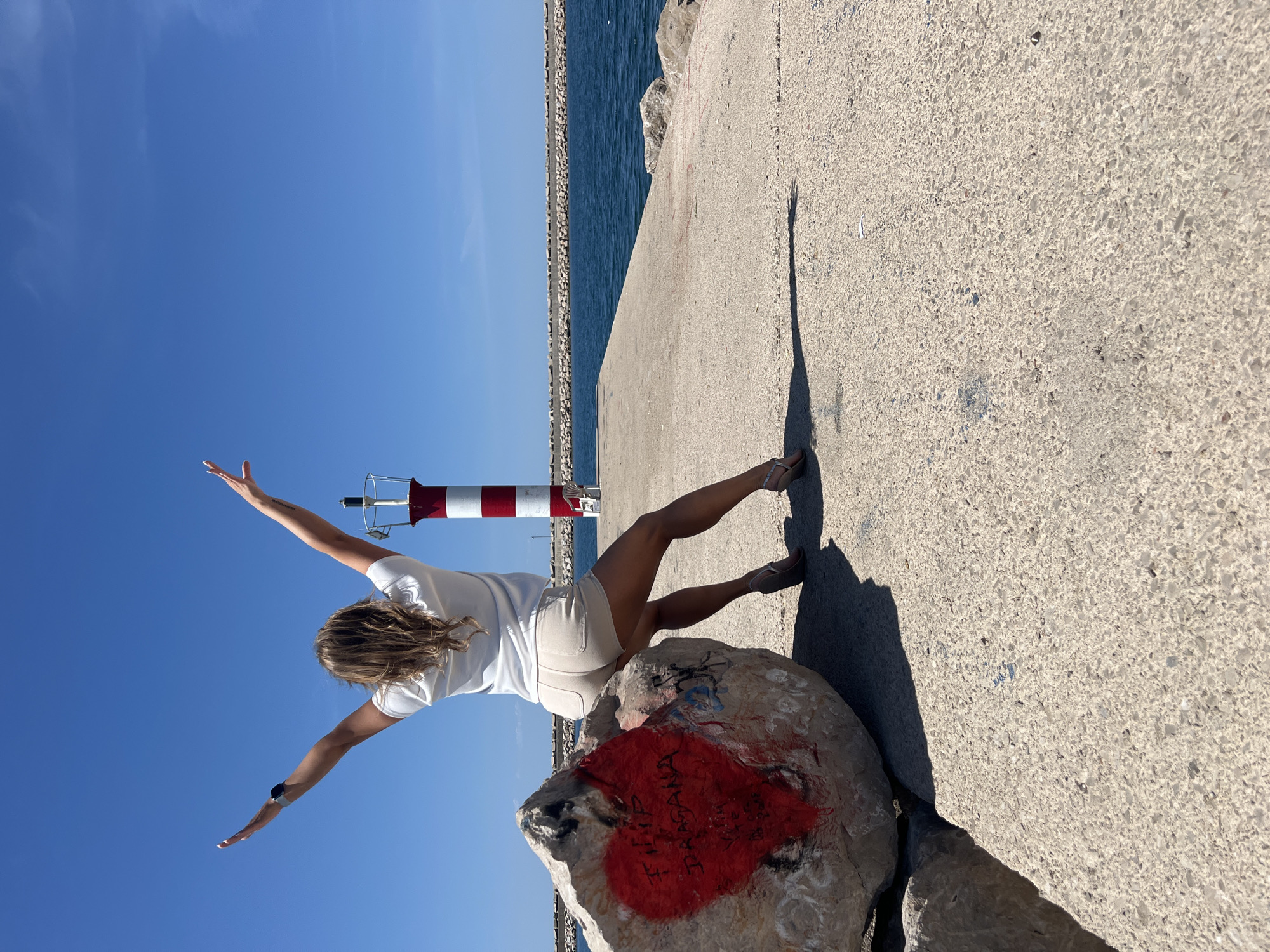 A full-body shot captures a woman from behind, posing on a sunny concrete pier in Bar, Montenegro. She is leaning against a large rock, with her arms outstretched sideways and her head turned towards the vast blue sea on the left. She wears a white t-shirt, light-colored shorts, and sandals. Behind her, on the pier, stands a small red and white striped navigational beacon or lighthouse.
The scene depicts a clear, bright day with intense sunlight, evidenced by the strong shadows cast on the concrete, including the woman's elongated shadow. The sea extends to the horizon, bordered by a long, rocky breakwater. The sky is a vivid, cloudless blue.
A notable detail is the large rock the woman is leaning on, which has a red heart painted on its side. Inside the heart, some text is visible, appearing to read "FILIP" and "DRAGANA". The setting evokes a sense of openness and coastal tranquility.
