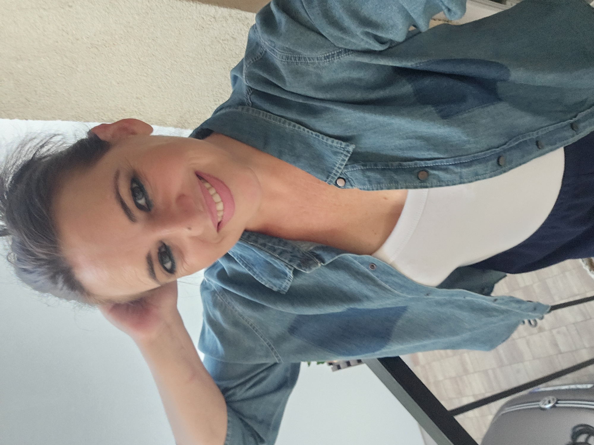 A woman is captured in a selfie-style shot, holding her hand to her head. She is wearing a denim shirt over a white tank top and dark bottoms. Her makeup is visible, including dark eyeliner and pink lipstick. The background appears to be an outdoor setting with a textured wall and a railing. The scene suggests a casual moment, possibly on a balcony or terrace. The location in Hévíz, Hungary, adds context to the geographical setting of the image, implying the photo was taken in this specific Hungarian city.