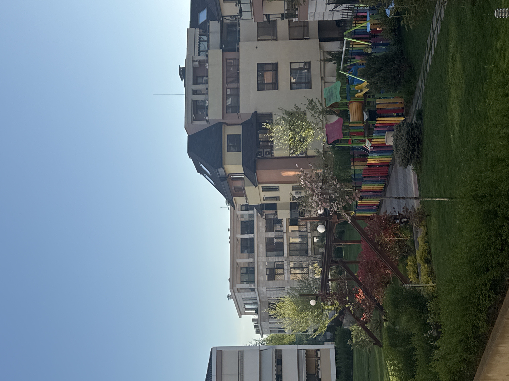 The image shows a residential area in Sofia, Bulgaria, featuring apartment buildings and a playground. The buildings are multi-story structures with a mix of architectural styles, including flat roofs and angled roofs. The exteriors are painted in light shades with dark window frames. A playground with colorful play equipment is situated near the buildings. The area includes green spaces with trees and grass. The sky is clear and blue.