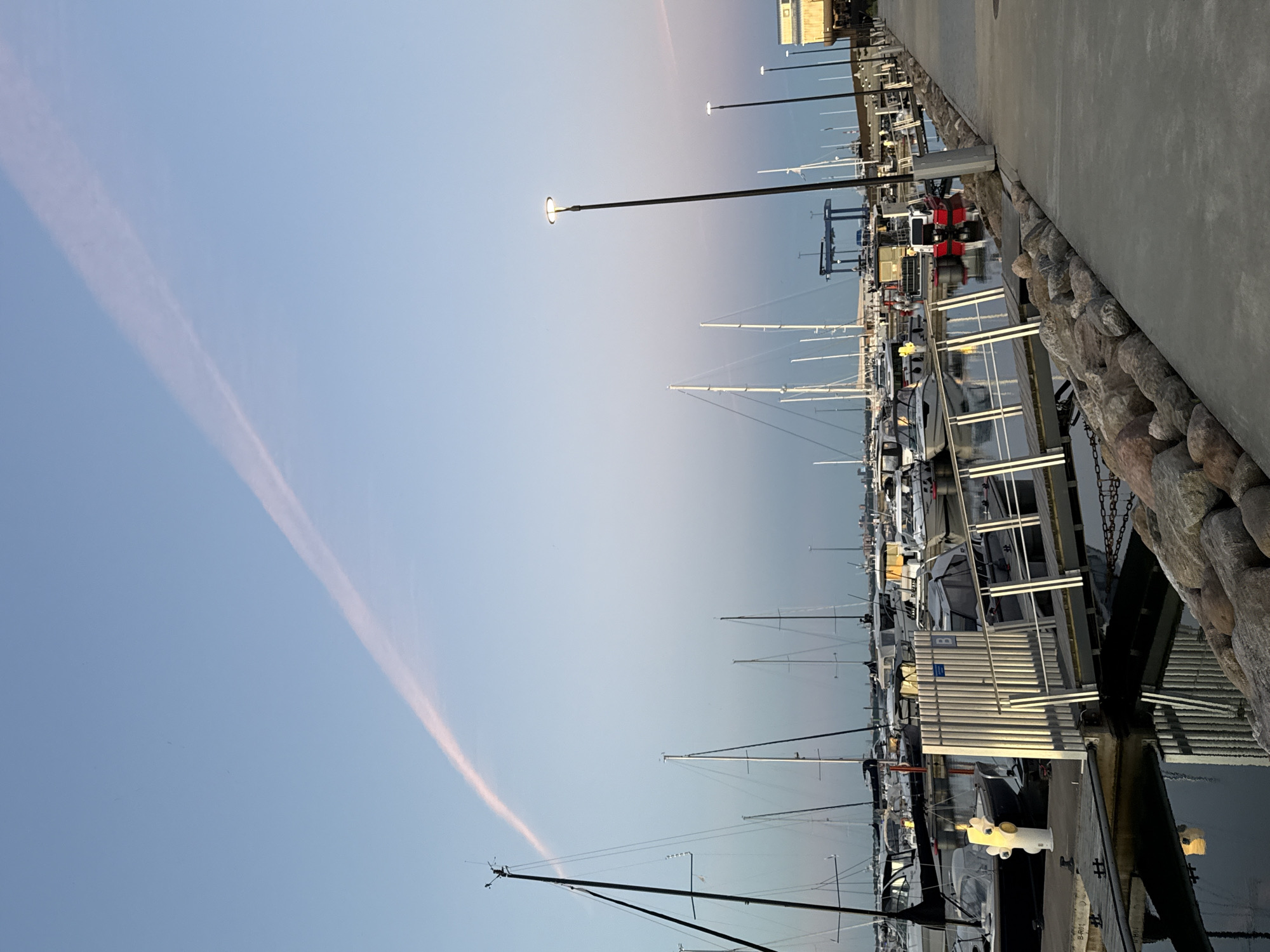 This is a wide-angle, eye-level photograph taken from a concrete pathway along a marina, likely in the city of Tallinn, Estonia, given the architectural style of the distant buildings and the presence of a prominent cargo crane which is typical of port areas. The image captures a serene scene at dusk or dawn, with a clear sky displaying a subtle gradient of light blue and pink from the setting or rising sun, intersected by a faint, pink contrail.
In the foreground, a paved walkway runs diagonally from the bottom right corner towards the center of the image, bordered by a line of large, rough-hewn rocks. A metal railing runs along the edge of the walkway, overlooking the water.
The marina itself is filled with numerous sailboats and motorboats docked side-by-side. Many of the sailboats have tall, prominent masts. The water in the marina is calm, reflecting the lights from the boats and the surrounding structures. A gangway with a metal railing extends from the pathway onto the docks, allowing access to the boats. A distinctive yellow fire hydrant is visible near the gangway.
Streetlights, turned on due to the low light conditions, are positioned along the pathway and the marina. In the background, there are more boats, the outline of a large industrial crane, and a building with a light-colored facade. The overall atmosphere is peaceful and quiet, suggesting an early morning or late evening moment. No people are clearly visible in the frame.