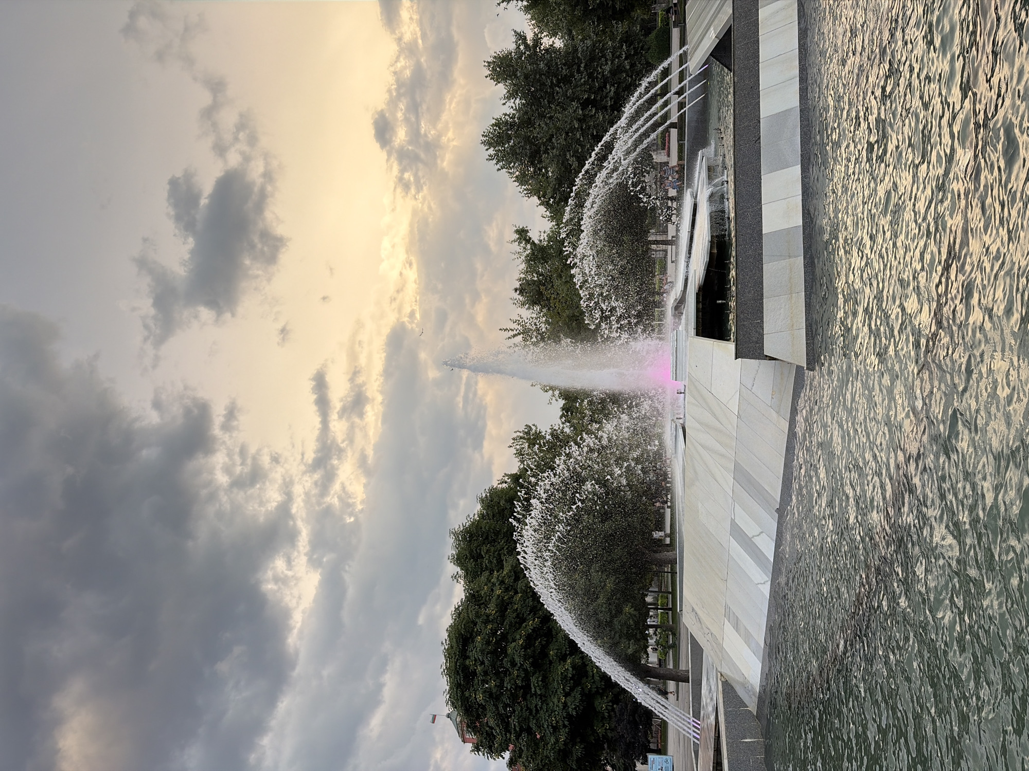 A wide-angle, eye-level shot captures a monumental fountain in the city of Pleven, Bulgaria. The fountain, constructed from white marble with dark grey accents, dominates the foreground with its geometric tiers and cascading water. A central jet shoots upwards, illuminated by pink lights at its base, while smaller streams fan outwards, creating a veil of mist. Beyond the fountain, lush green trees line a park area, their branches extending towards the sky. In the background, a red building with a Bulgarian flag flying on top stands prominently, suggesting a civic or administrative location. The sky above is a canvas of dramatic clouds, transitioning from grey at the top to a soft yellow and orange hue near the horizon, hinting at either sunrise or sunset. The water in the foreground ripples gently, reflecting the muted light and the surrounding environment.