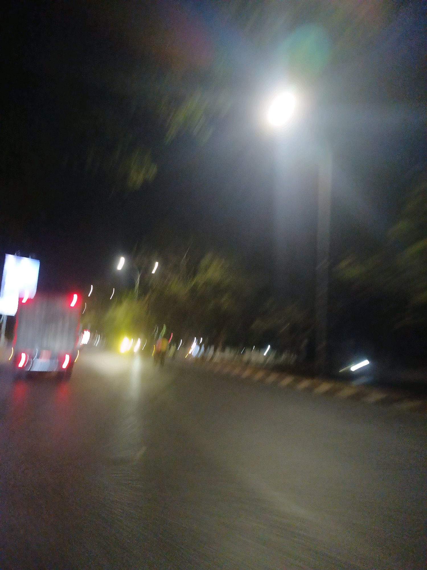 This is a blurred, nighttime photograph taken from a moving vehicle on a wet road in Makassar, Indonesia. The image shows a street scene with several vehicles and streetlights. On the left side of the road, a truck with its red taillights illuminated is visible. Further down the road, other vehicles and streetlights create streaks of light due to motion blur. Trees line the right side of the road, and the overall atmosphere suggests a humid, possibly rainy evening.