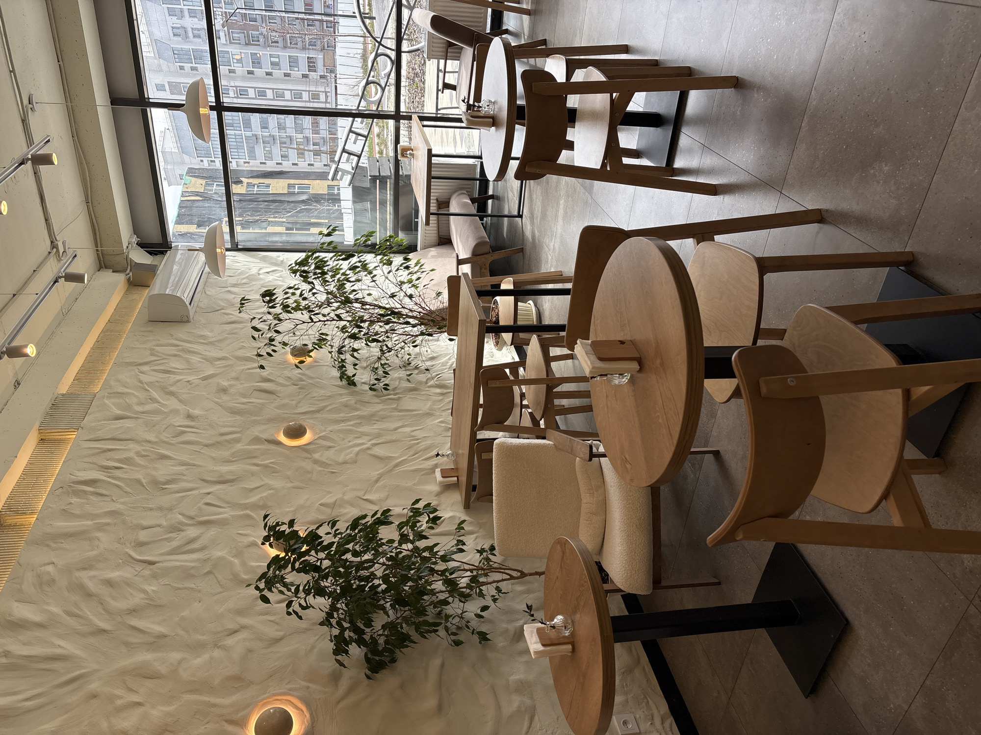 The image shows the interior of a cafe or restaurant in Ulyanovsk, Russia. The room has a modern design with a textured white wall, wooden tables and chairs, and large windows overlooking a cityscape with multi-story buildings. Potted plants are placed along the wall.