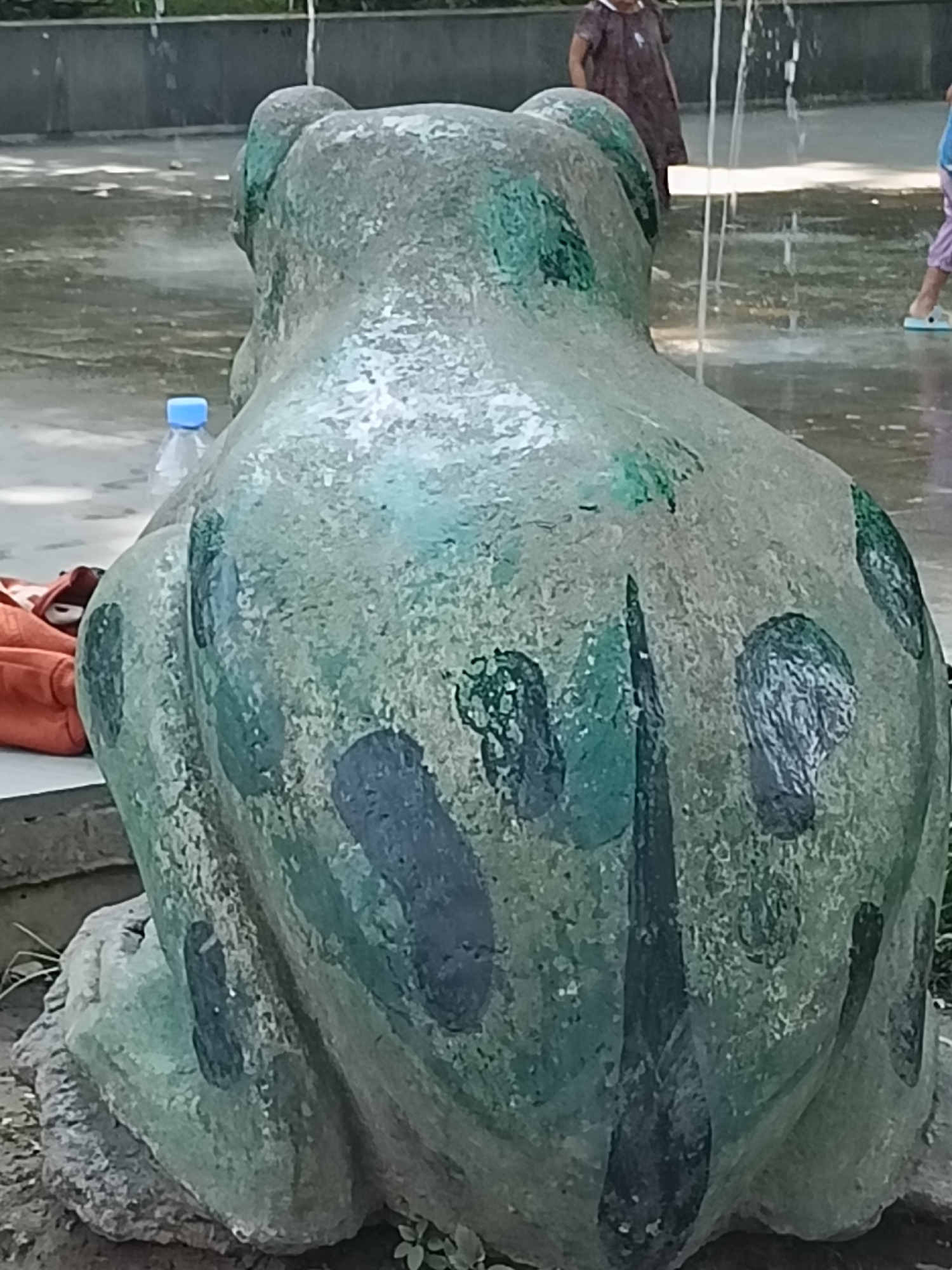 An outdoor scene features a large, weathered, green-painted concrete frog statue. The statue is positioned from a rear perspective, facing away from the viewer. The frog's back is textured and shows patches of darker green or black paint, possibly representing camouflage or natural markings. The statue sits on a rocky base in what appears to be a park or public space. In the background, water from a fountain is visible, creating a misty effect and reflecting light on the wet ground. A plastic water bottle with a blue cap is placed near the statue. Further in the background, a person wearing a dark patterned dress is partially visible, and another person's legs wearing light-colored pants and blue sandals are also seen, suggesting the presence of other visitors. The overall environment suggests a warm, possibly sunny day.