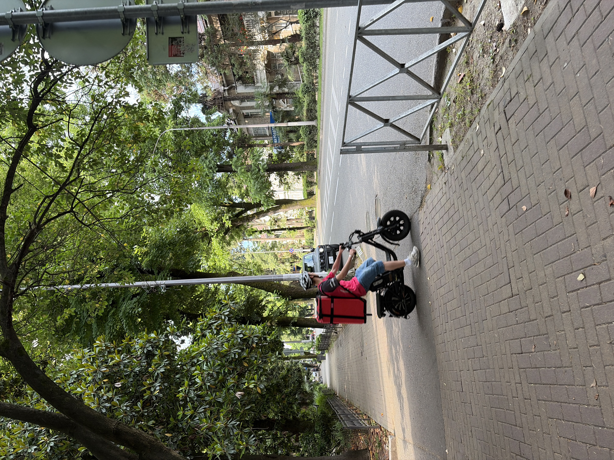 A person wearing a helmet and a pink shirt rides an electric scooter along a brick sidewalk in Sochi, Russia. The scooter has a large red insulated bag attached to the back. The sidewalk is lined with trees and a metal fence separates it from a paved road. Buildings are visible across the street, and a black SUV is parked on the side of the road. The sun is shining, casting shadows on the sidewalk.