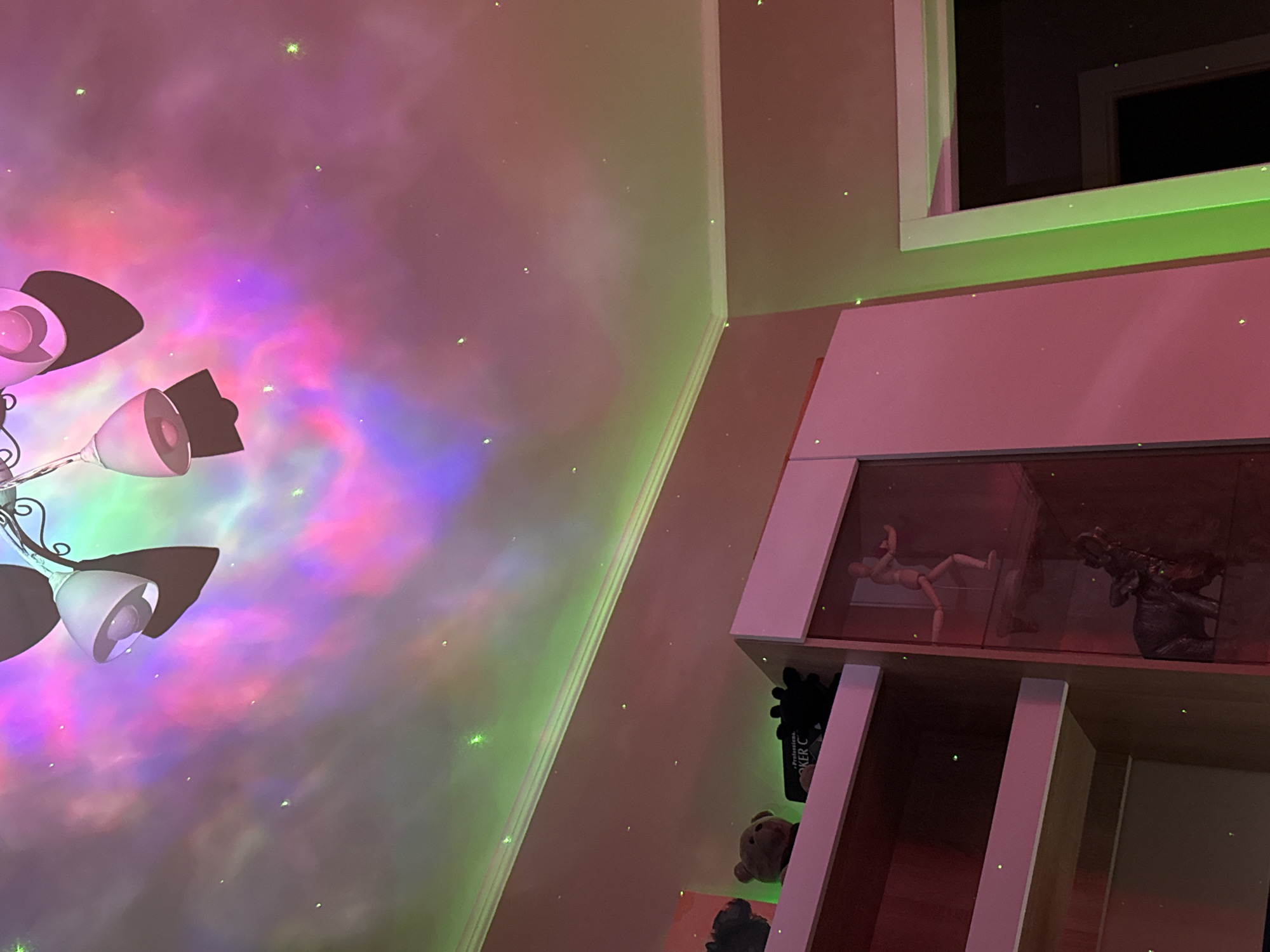 The image shows the interior of a room with a lamp illuminating the wall with a colorful, nebula-like pattern. The light source is positioned on the lower left. The wall is also lit with small green points of light, possibly from a laser projector. To the right of the wall is a multi-tiered shelf or display case containing objects, including a mannequin and a dragon figurine in the upper section, and a book titled "Poker C" in the shelf beneath. At the top right, a window frame is visible. The room appears to be indoors, possibly a bedroom or playroom. There is no indication of a specific city or location.