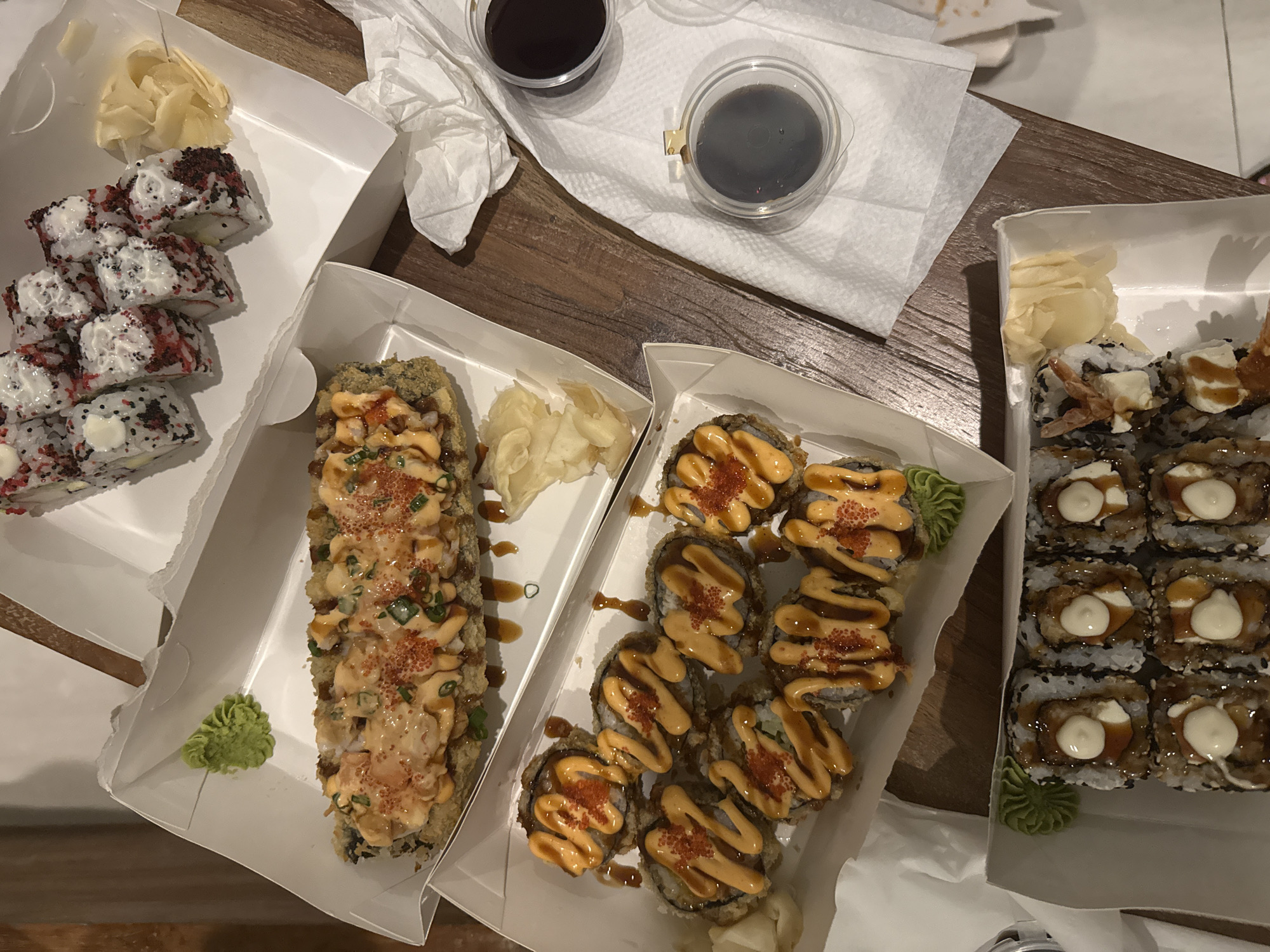 A top-down view displays an assortment of sushi in white takeout containers, laid out on a light brown wooden table. The scene suggests a meal, likely takeout or delivery.
In the upper left container, several maki rolls are visible, topped with a white creamy substance, possibly cream cheese or mayonnaise, and sprinkled with red and black specks, resembling masago or another type of roe. A portion of thinly sliced pickled ginger rests beside the rolls.
Below this, a longer, deep-fried sushi roll takes center stage in another container. It is lavishly drizzled with an orange-pink sauce, garnished with chopped green onions, and speckled with red roe. A small mound of green wasabi paste is placed to its left.
To the right, a third container holds several smaller, individual deep-fried sushi pieces, each also topped with the same orange-pink sauce and red roe.
In the bottom right corner, the fourth container showcases sushi rolls characterized by black sesame seeds on the rice, a dark, glossy sauce (possibly eel sauce), and a distinct dollop of white creamy topping on each piece. Another swirl of green wasabi is present in this box.
Two small, clear plastic cups, filled with a dark liquid, likely soy sauce, are positioned on a white paper napkin in the upper central area of the frame. Additional crumpled white napkins are scattered nearby. The lighting suggests an indoor setting. No specific time of day or emotions can be inferred from the image. There is no visible text on any of the containers or items. While provided with a location context of "Ubud District, Indonesia," the image itself contains no explicit visual cues to confirm this geographical detail.
