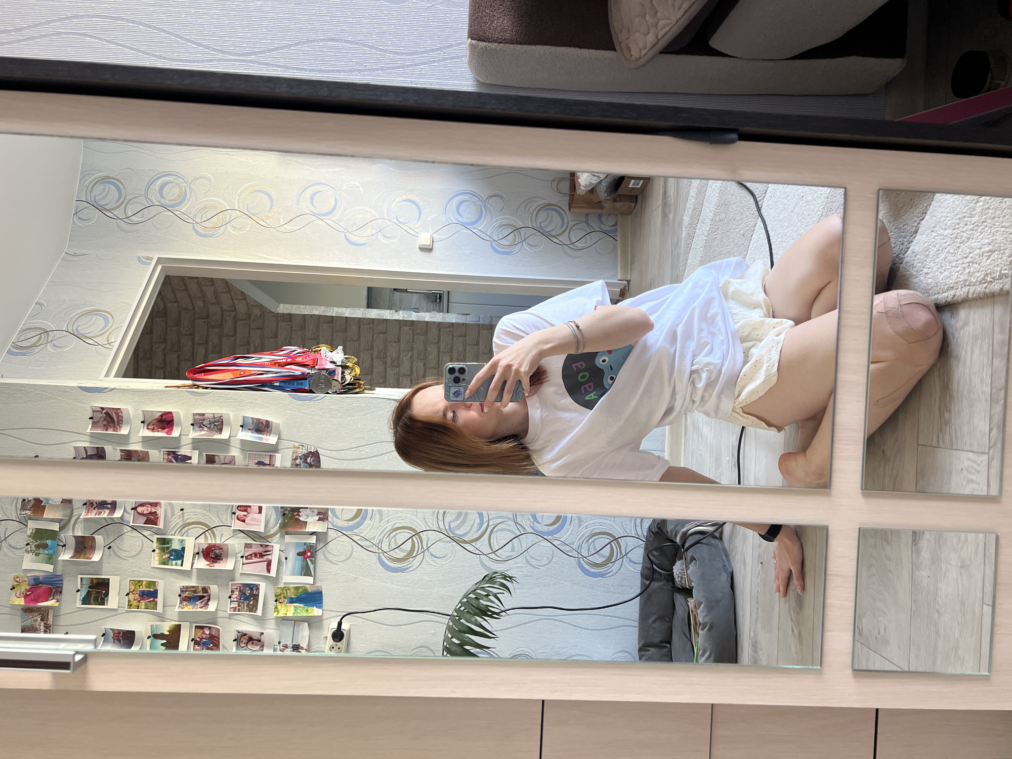 A young adult woman is reflected in a multi-paneled mirror in what appears to be an apartment or home in Chaikovsky, Russia. The woman is kneeling on a light-colored rug and holding a smartphone in her left hand to take a selfie in the mirror. She is wearing a white t-shirt and light-colored shorts. Her knees have bandages on them.
The mirror reflects a room with patterned wallpaper. On the wall to the left, multiple small photographs are displayed with wire lights around them. In the background of the mirror, there is an open doorway leading to a room with brick-style walls.
The apartment interior is simple. The furniture includes a pet bed and storage unit. A potted plant can be seen reflecting in the mirror.