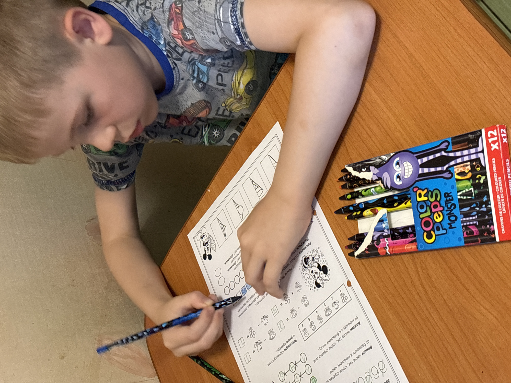 A boy sits at a wooden desk working on a worksheet. He is wearing a gray shirt with cartoon images. A pack of "Color Peps Monster" colored pencils rests to his right. The worksheet appears to contain a matching activity and simple math problems. The location, Rusanovo, Russia, cannot be verified from the image.