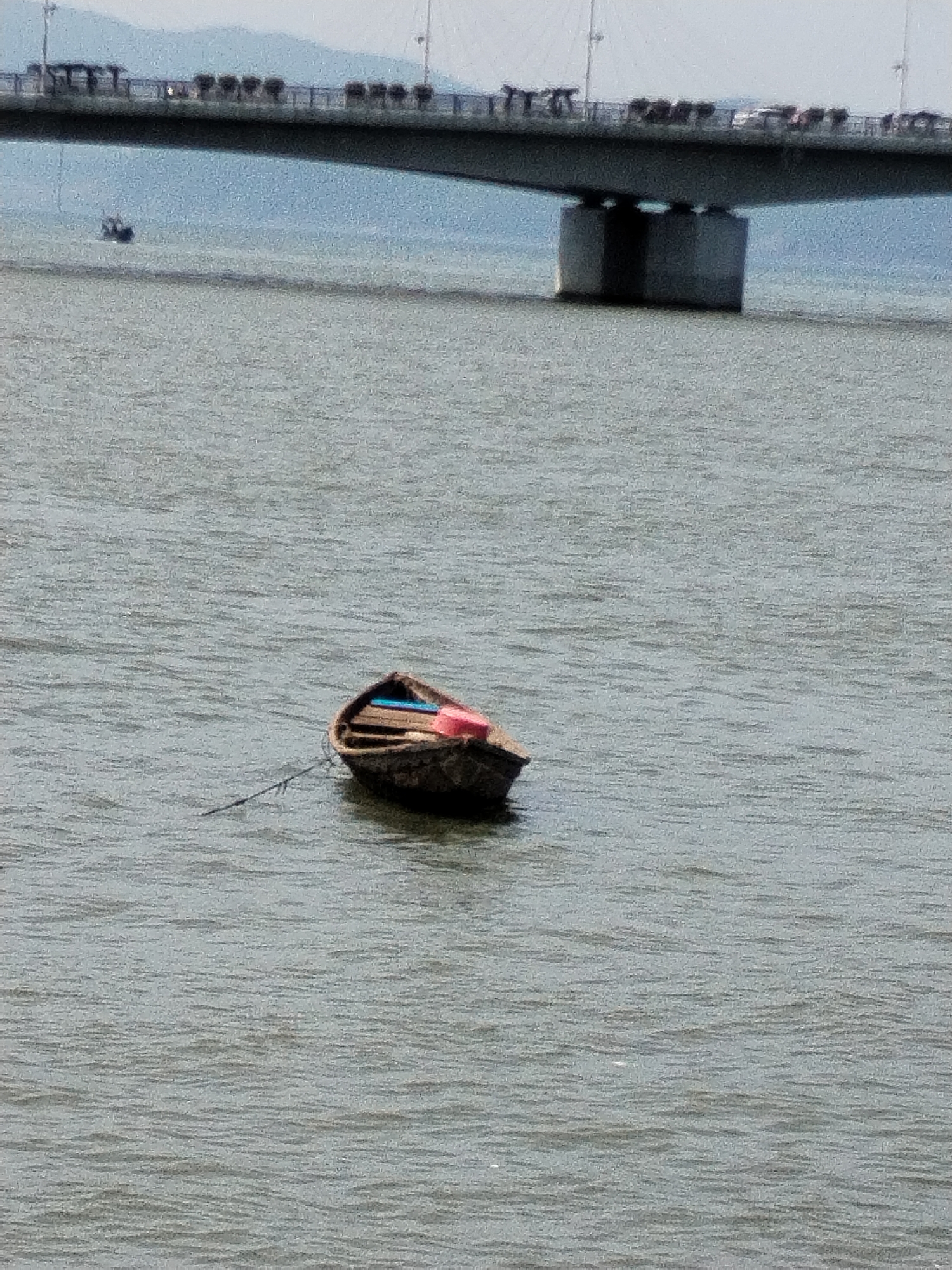A single, weathered wooden boat with red and blue accents floats on calm, grey water. The boat is moored by a rope that trails off to the left. In the background, a large concrete bridge spans the water, with a line of silhouetted vehicles visible on its deck. The sky is overcast and the water is a muted grey, suggesting a cloudy day. The scene appears to be from a city harbor or bay, possibly in Bac Nha Trang Ward, Vietnam, given the architectural style of the bridge and the type of vessel.