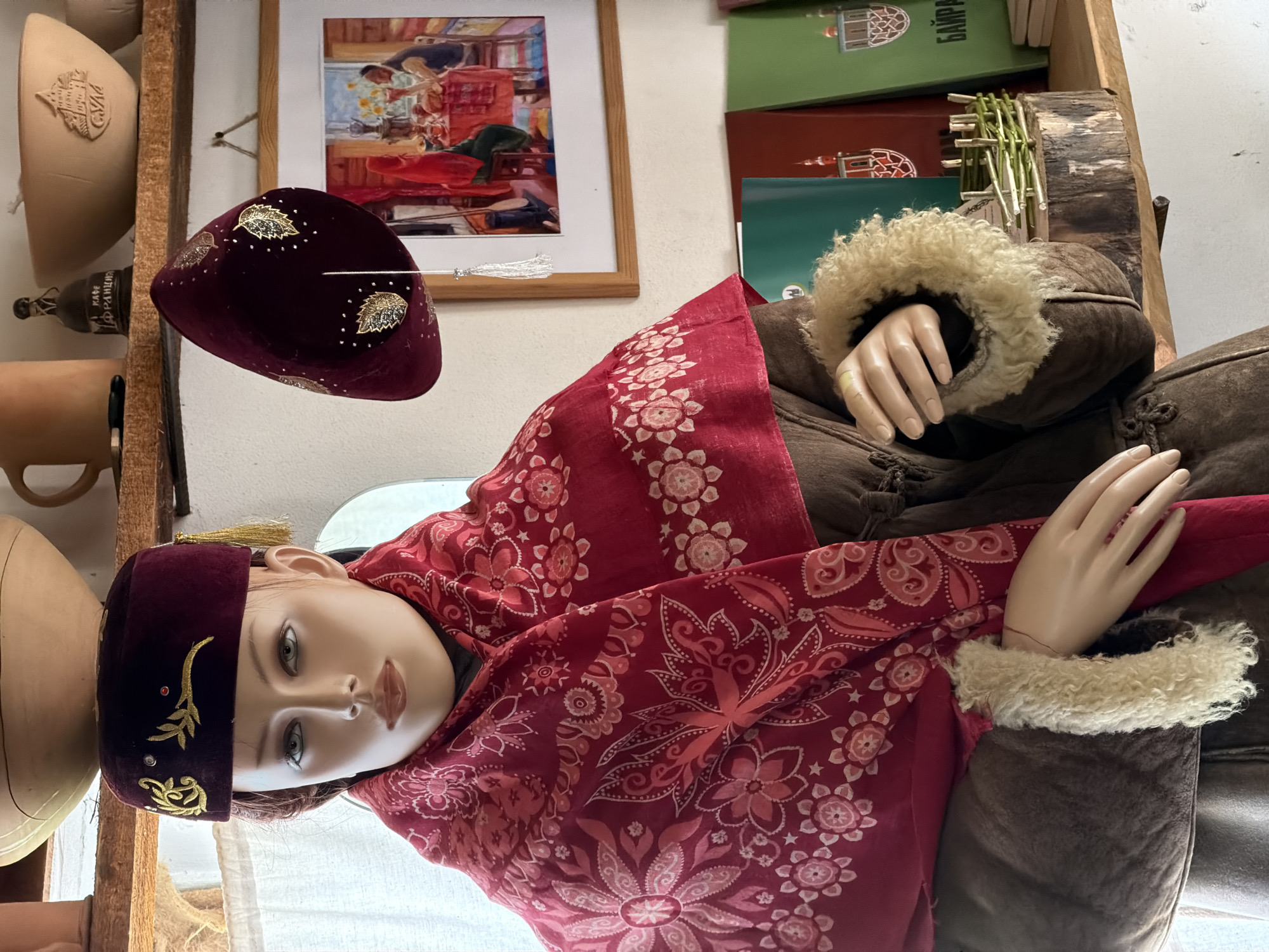The image displays a mannequin dressed in traditional attire, likely in a shop or museum. The mannequin wears a dark red velvet cap adorned with gold embroidery and a red patterned scarf. It is also dressed in a brown coat with fur trim around the cuffs. In the background, a framed painting depicts a domestic scene with people gathered around a table. Shelves behind the mannequin hold various ceramic objects, including bowls and mugs, and a collection of books or notebooks. A small woven decorative fence sits on a wooden log. The scene suggests an indoor setting with cultural artifacts and decorative elements. The location, Літвенскі сельскі Савет, Belarus, implies a setting within a rural administrative area in Belarus, potentially a cultural center, a craft shop, or a historical exhibition space.