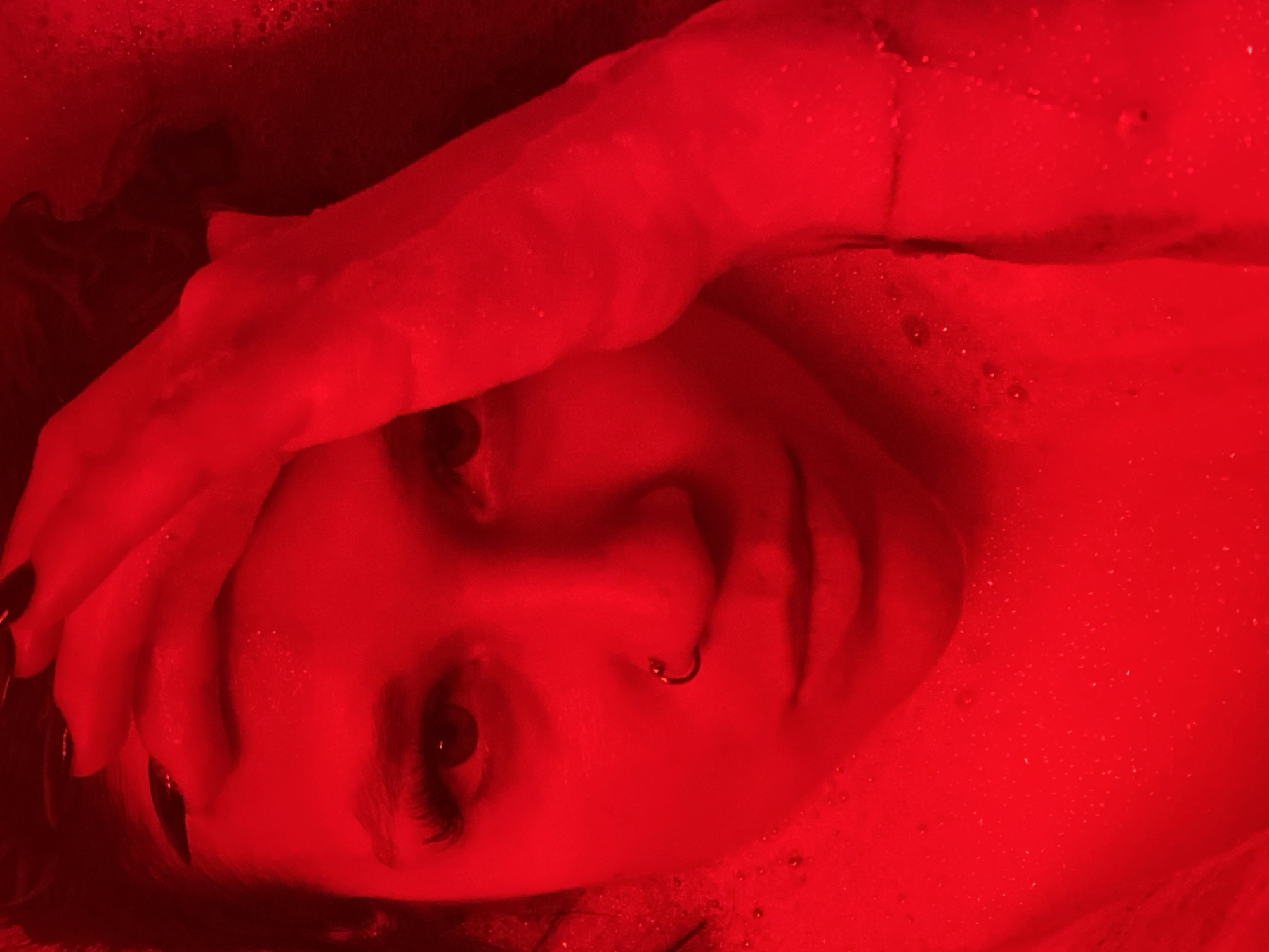 A close-up, top-down shot of a person's face illuminated by red light. The person has dark nail polish and a nose ring. Their right hand is raised to their forehead. Bubbles are visible in the water surrounding them. The image is a selfie taken in Chaikovsky, Russia.