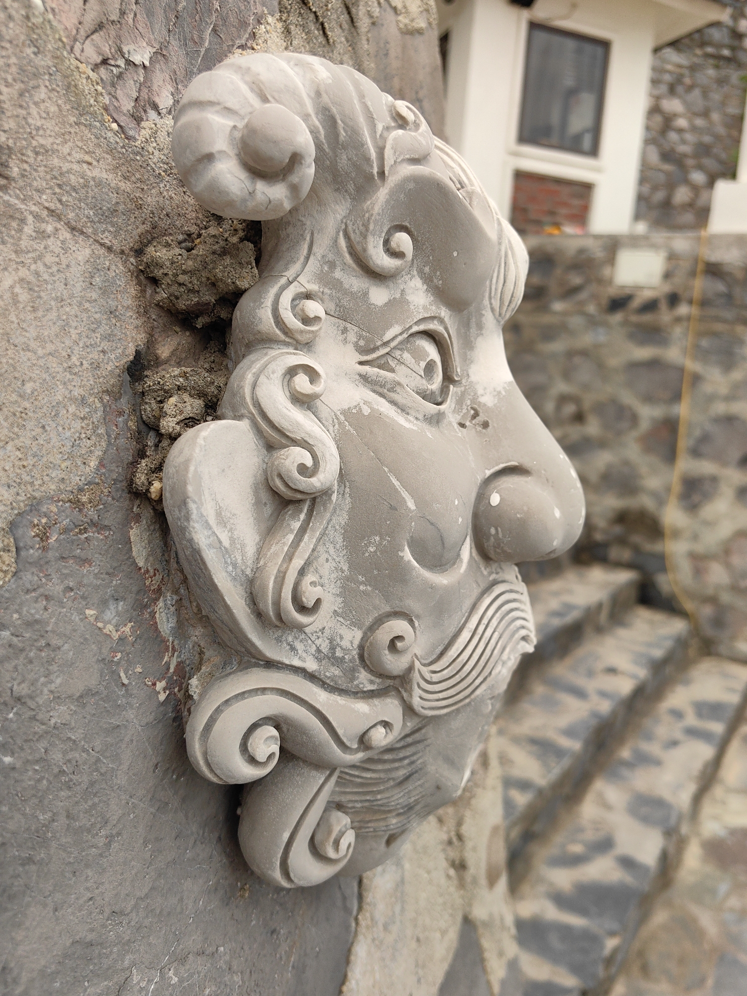 A close-up shot shows a decorative stone carving of a human face attached to a rough, textured stone wall. The face features stylized swirls and curls along its periphery, a prominent nose, and an eye. The backdrop reveals stone stairs and a building with a window. The location is Hải Phòng, Vietnam.